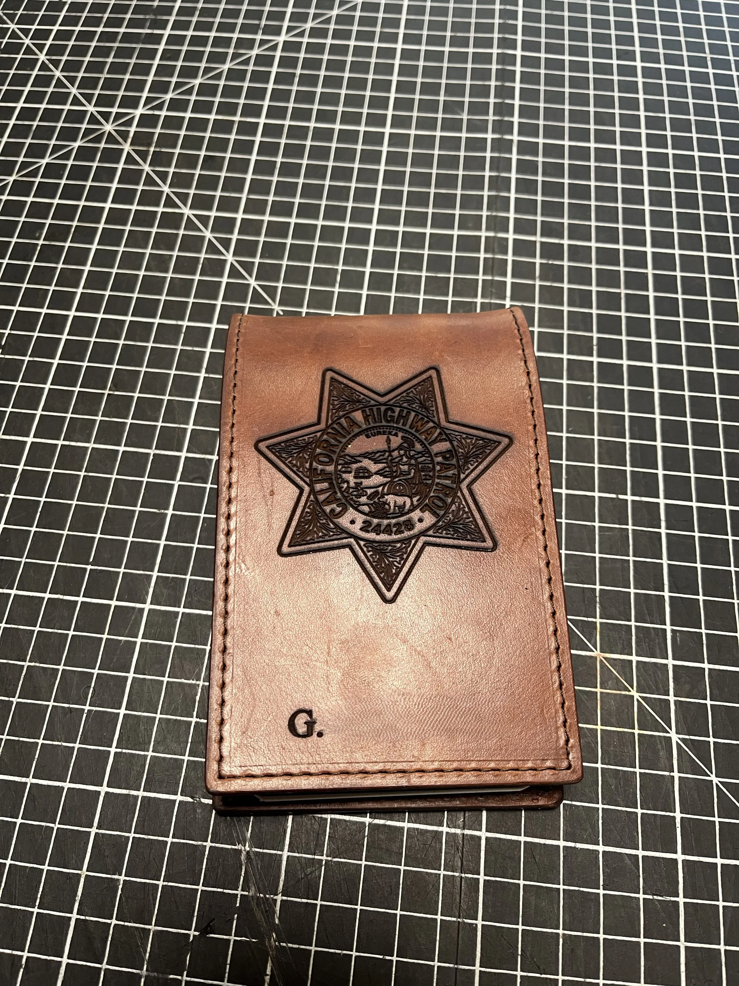 A brown leather wallet with the emblem of a police badge embossed on it, placed on a black cutting mat with a grid pattern.