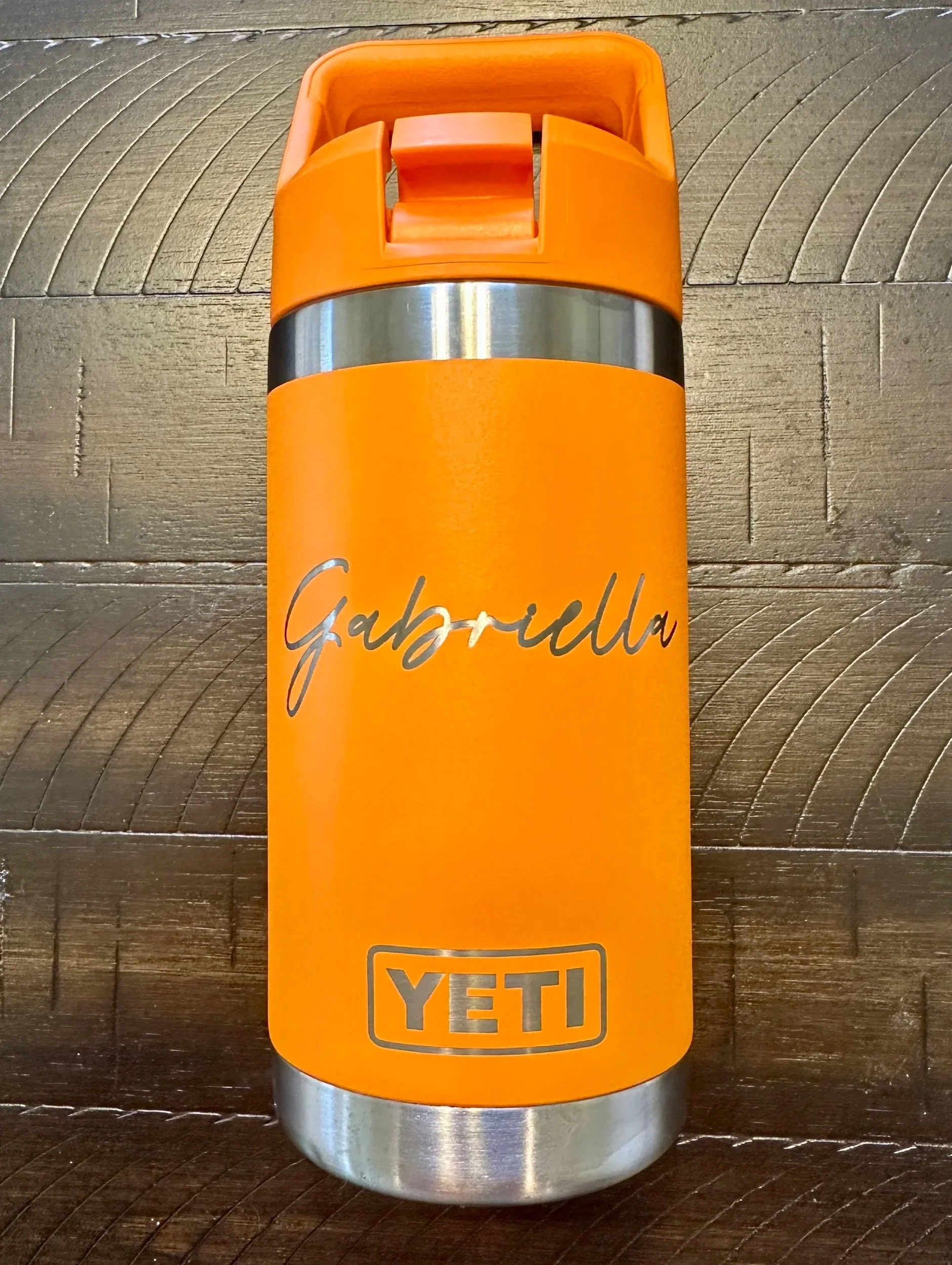 Close-up of an orange YETI Rambler bottle with a black stripe and the word "Gabriella" written in cursive, placed on a wooden surface.