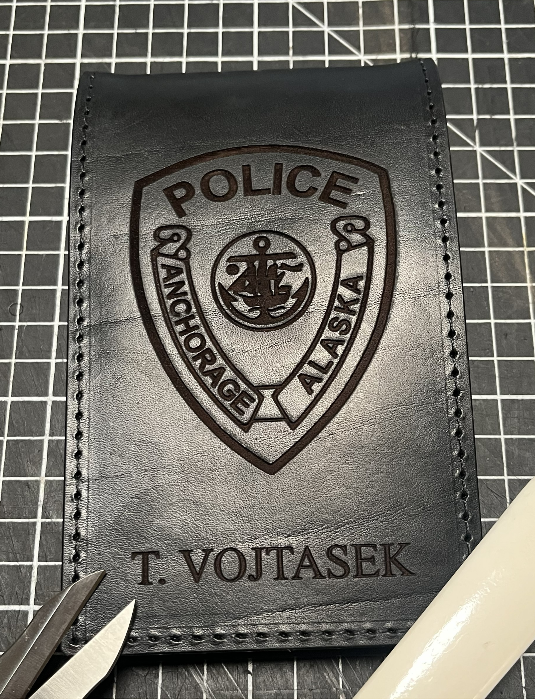 Black leather badge with embossed police emblem and text, placed on cutting mat. Tools include tweezers and a craft knife, indicating a craft or customization process.