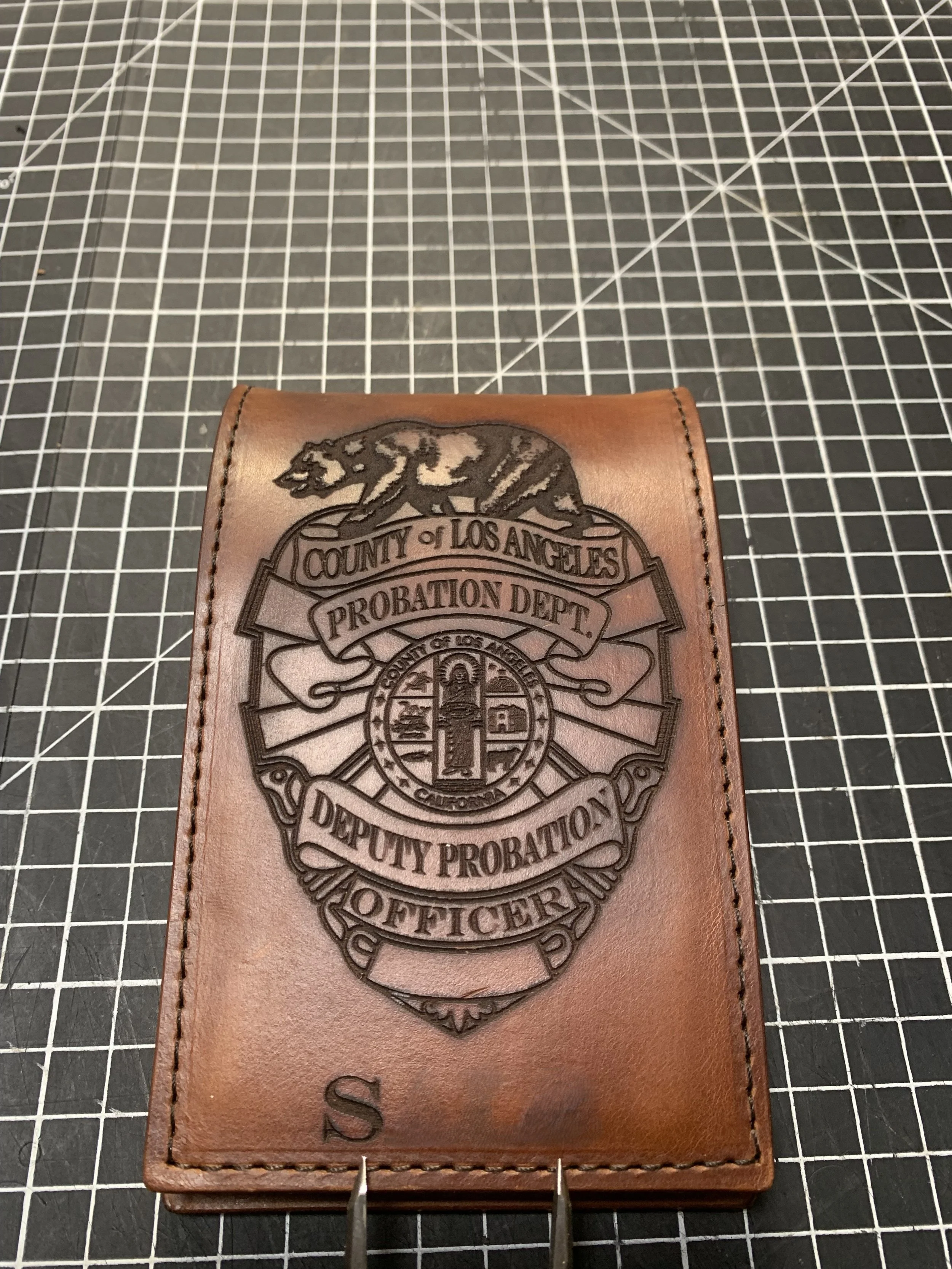 Leather badge or card holder with an embossed emblem and text indicating the County of Los Angeles Probation Department, Department Office, featuring a panther and a torch, on a black grid cutting mat.