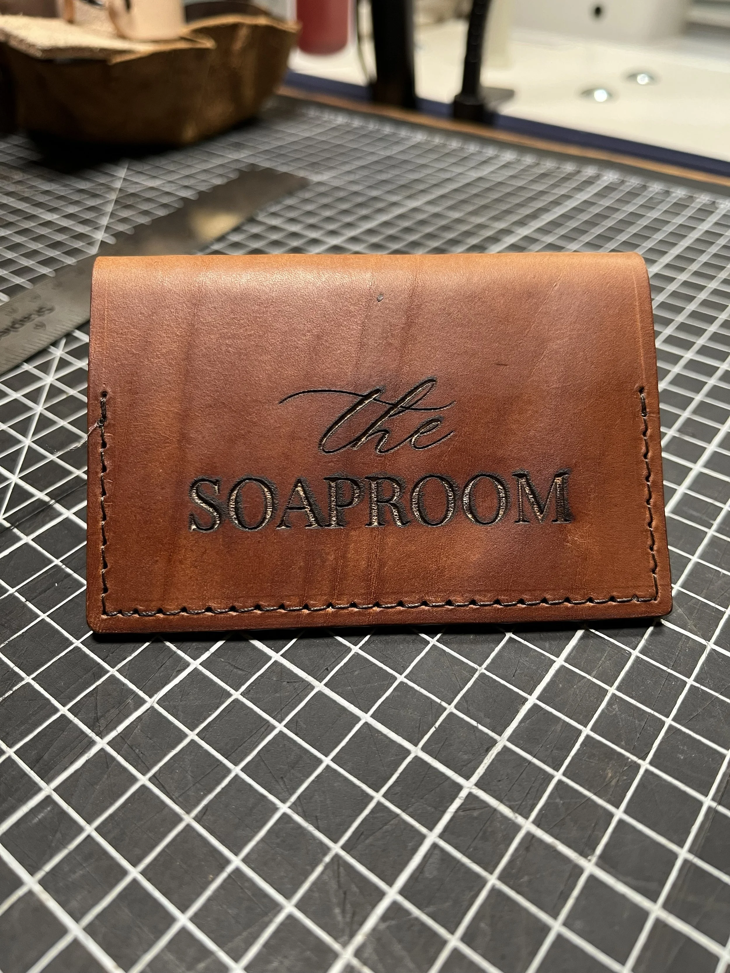 A brown leather cardholder with the text 'The Soaproom' embossed on it, placed on a metal wire grid surface.