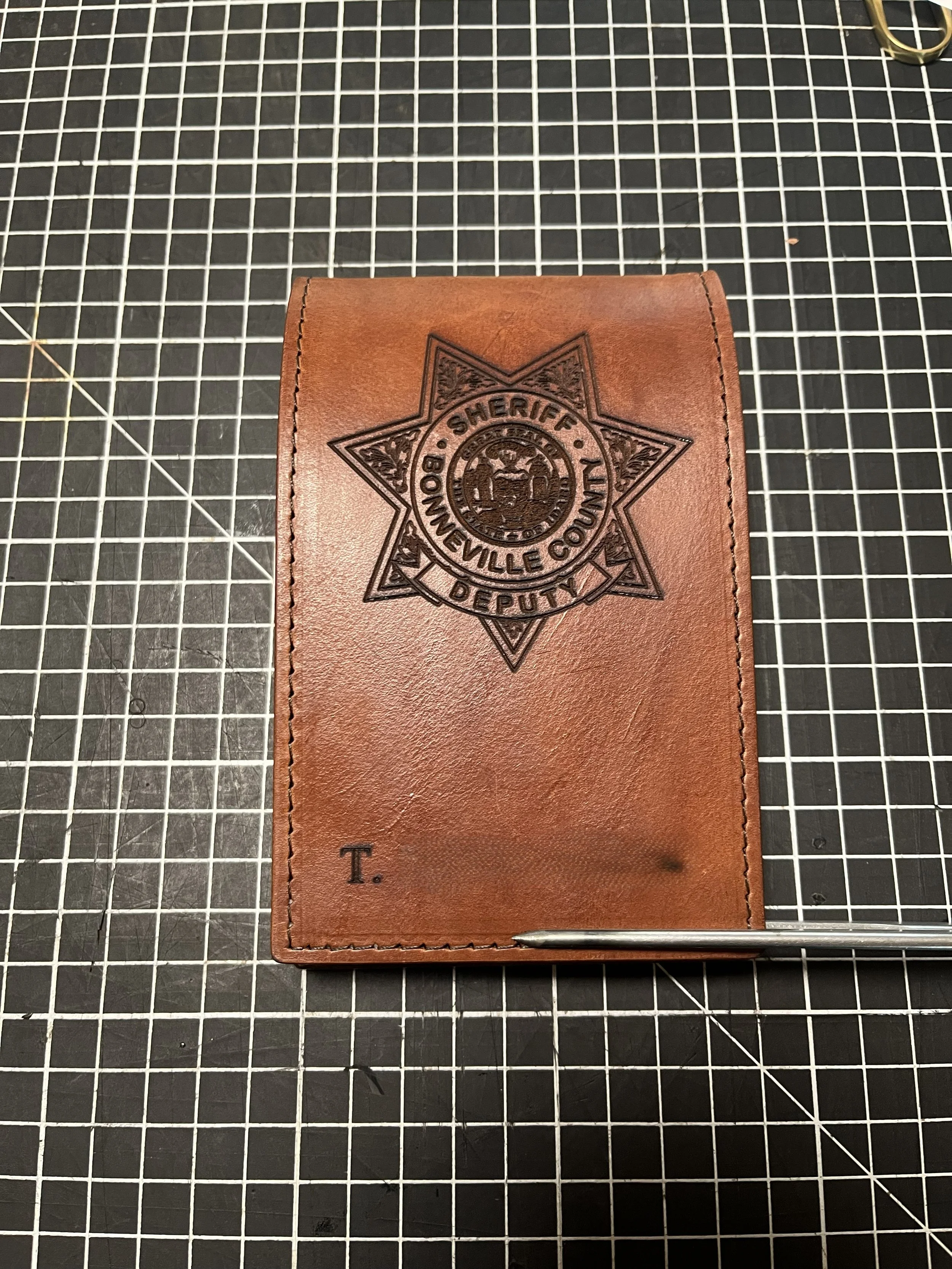 Brown leather wallet with a Sheriff's badge logo embossed on it, placed on a black cutting mat with grid lines, and a metallic pen at the bottom edge.