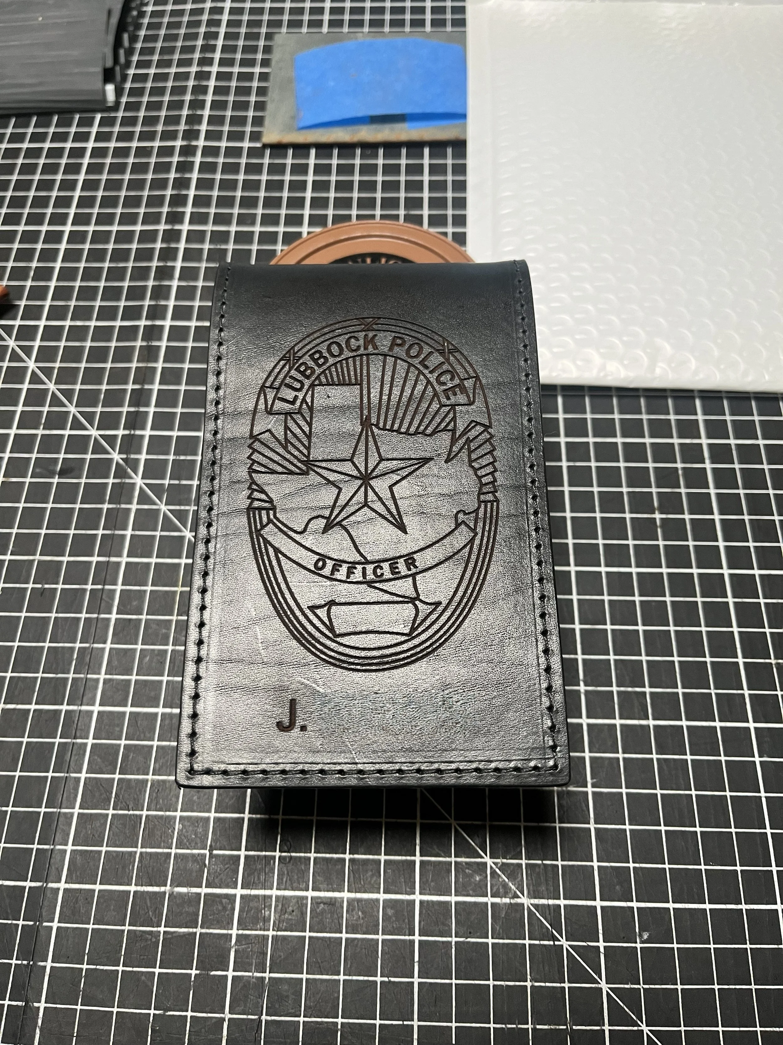 A black leather badge holder with the Lubbock Police badge embossed on it and the initial J at the bottom, placed on a black and white grid cutting mat.