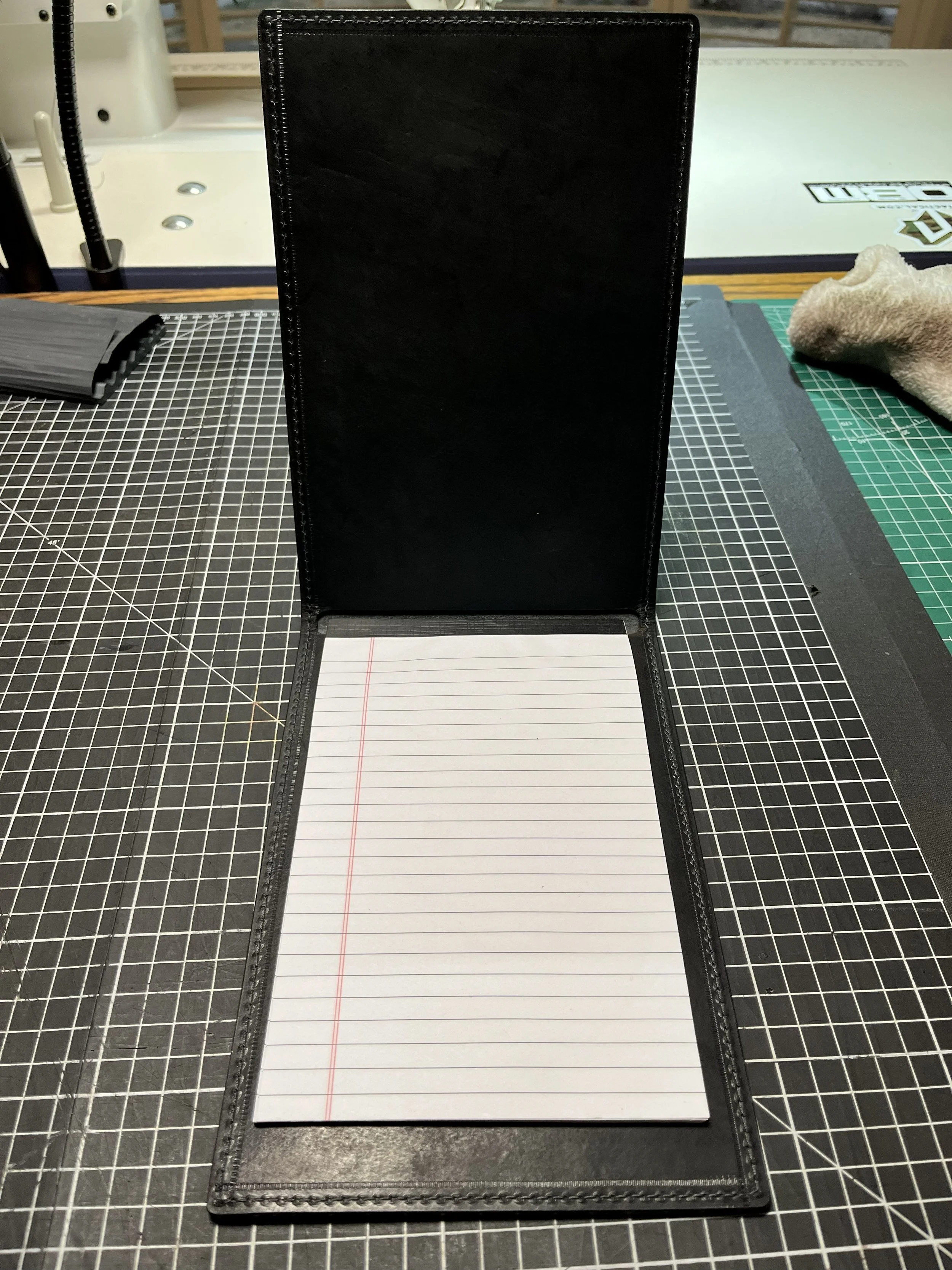 An open black leather portfolio with a notepad inside, placed on a black cutting mat with a grid pattern, on a worktable with tools and supplies in the background.
