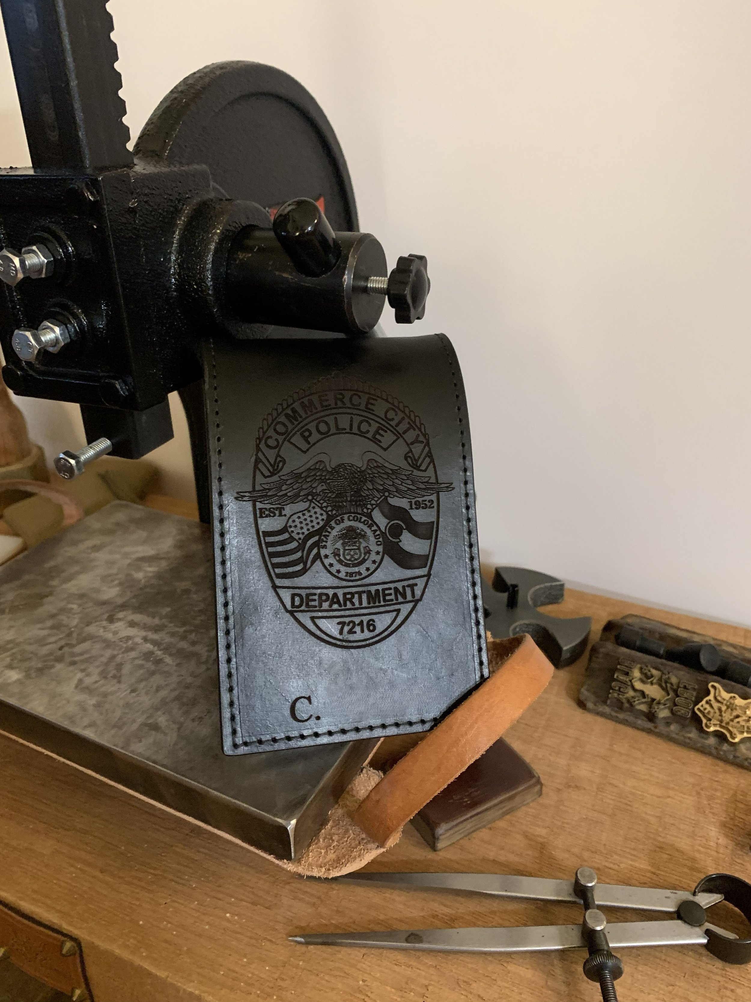 Close-up of a tool or device mounted on a workbench, with a badge from the Commerce City Police Department, Est. 1952, attached to it, surrounded by various small tools and objects, on a wooden surface.