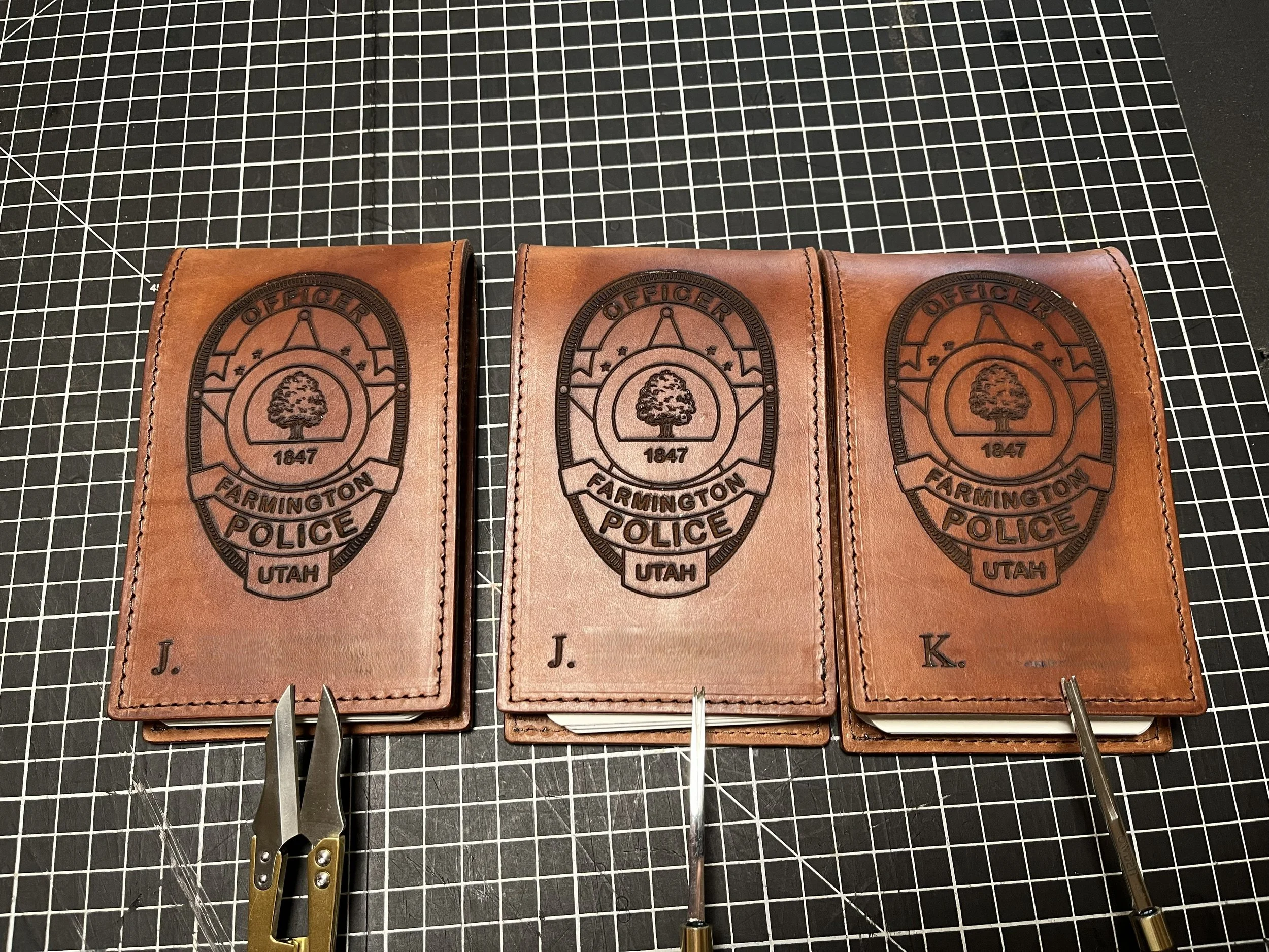Three leather pouches with a police badge emblem from Farmington, Utah, bearing the year 1847. The pouches are placed on a black grid work surface, with some jewelry-making tools in the foreground.