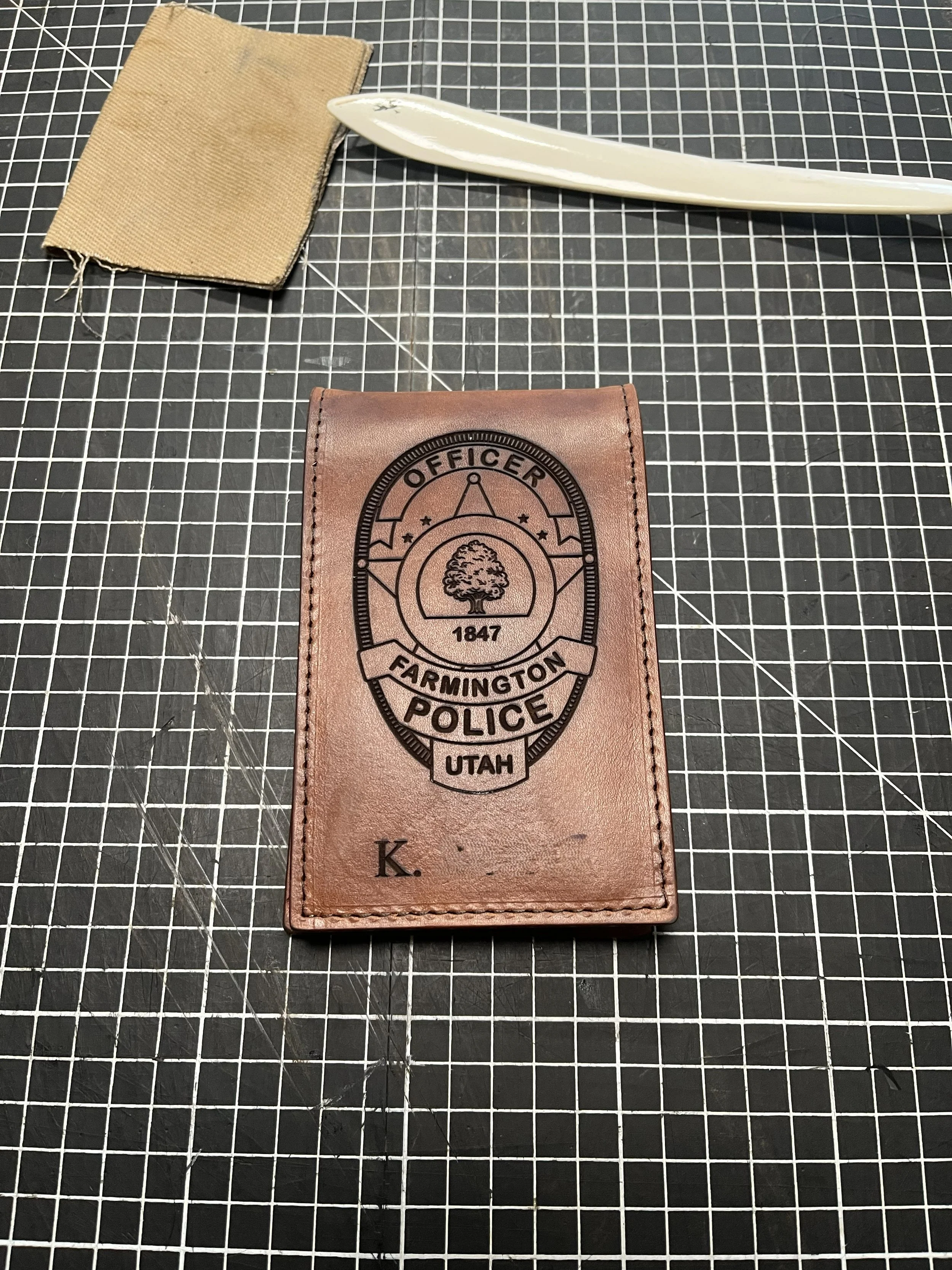 A leather card holder with a police badge logo from Farmington Police Department, Utah, placed on a cutting mat with a grid pattern. There is a beige fabric wallet, a white ceramic knife, and a clear plastic straw on the cutting mat.