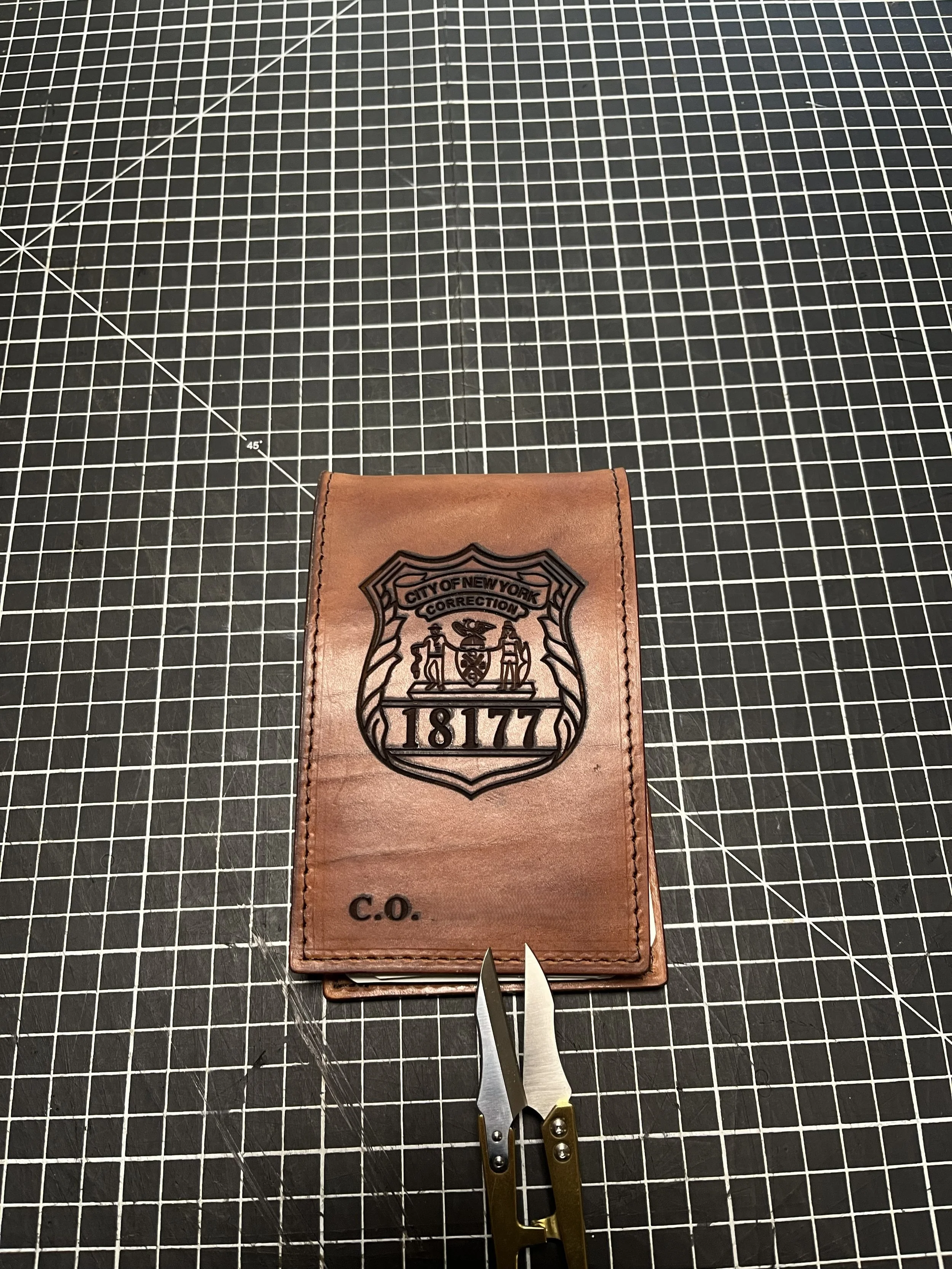 A brown leather wallet with a city of New York police badge logo and the text 'C.O.' on it, placed on a black grid cutting mat. There are scissors and a precision knife nearby.