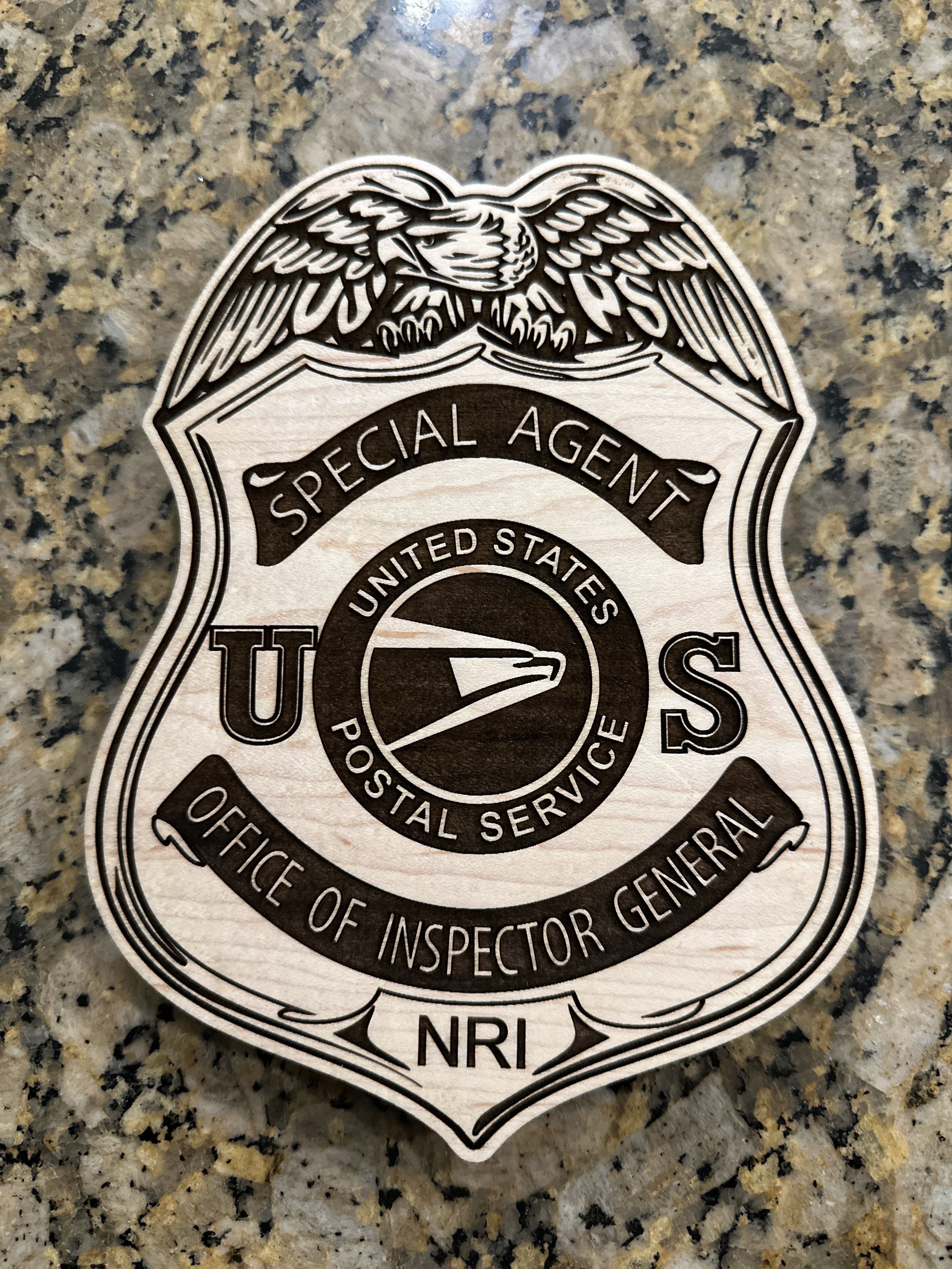 Close-up of a wooden badge with an engraving of an eagle at the top, containing text that reads 'Special Agent' and 'U.S. Postal Service' inside a circular emblem, resting on a speckled granite surface.