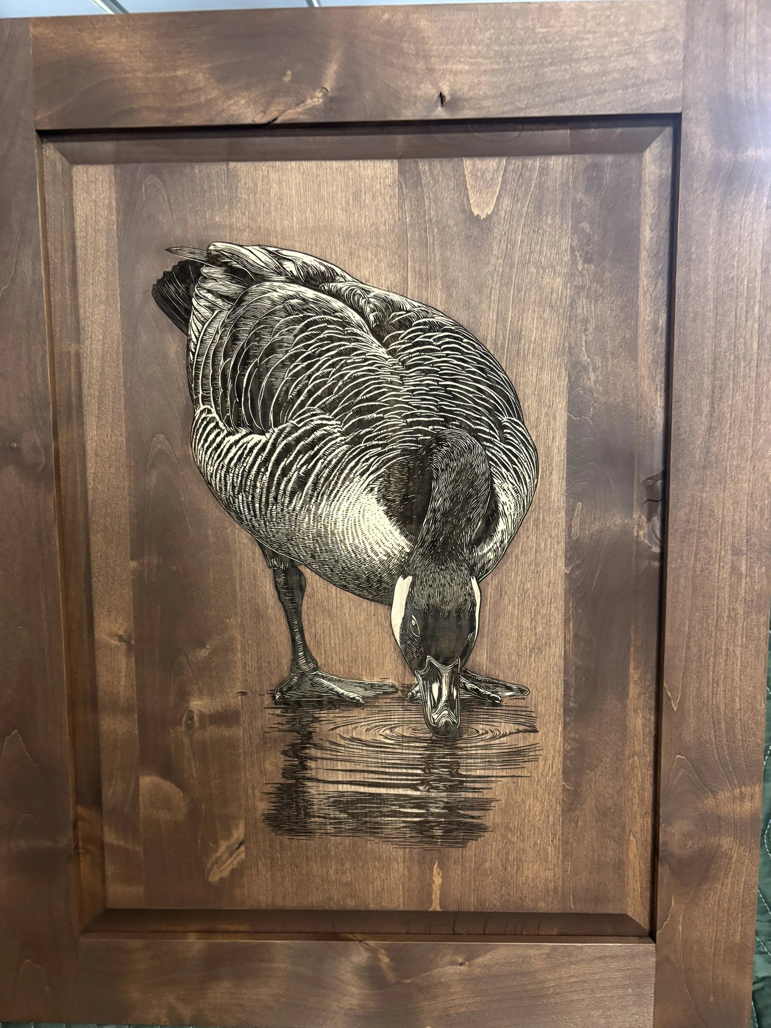 A black and white illustration of a duck standing on water, with its head bent down.