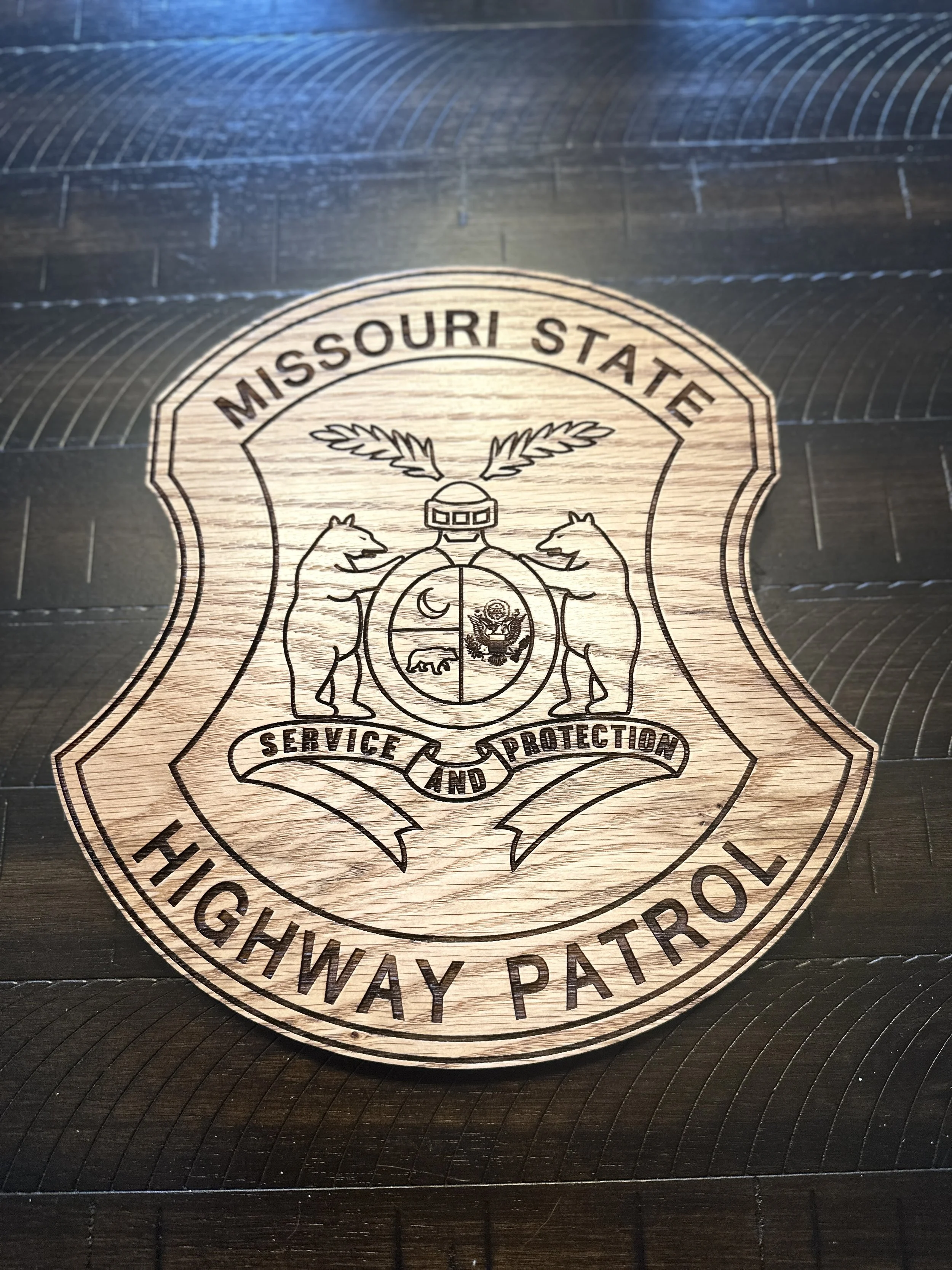 Emblem for Missouri State Highway Patrol engraved on a wooden surface, featuring a shield with a bear, a sword, and a motto reading 'Service and Protection'.