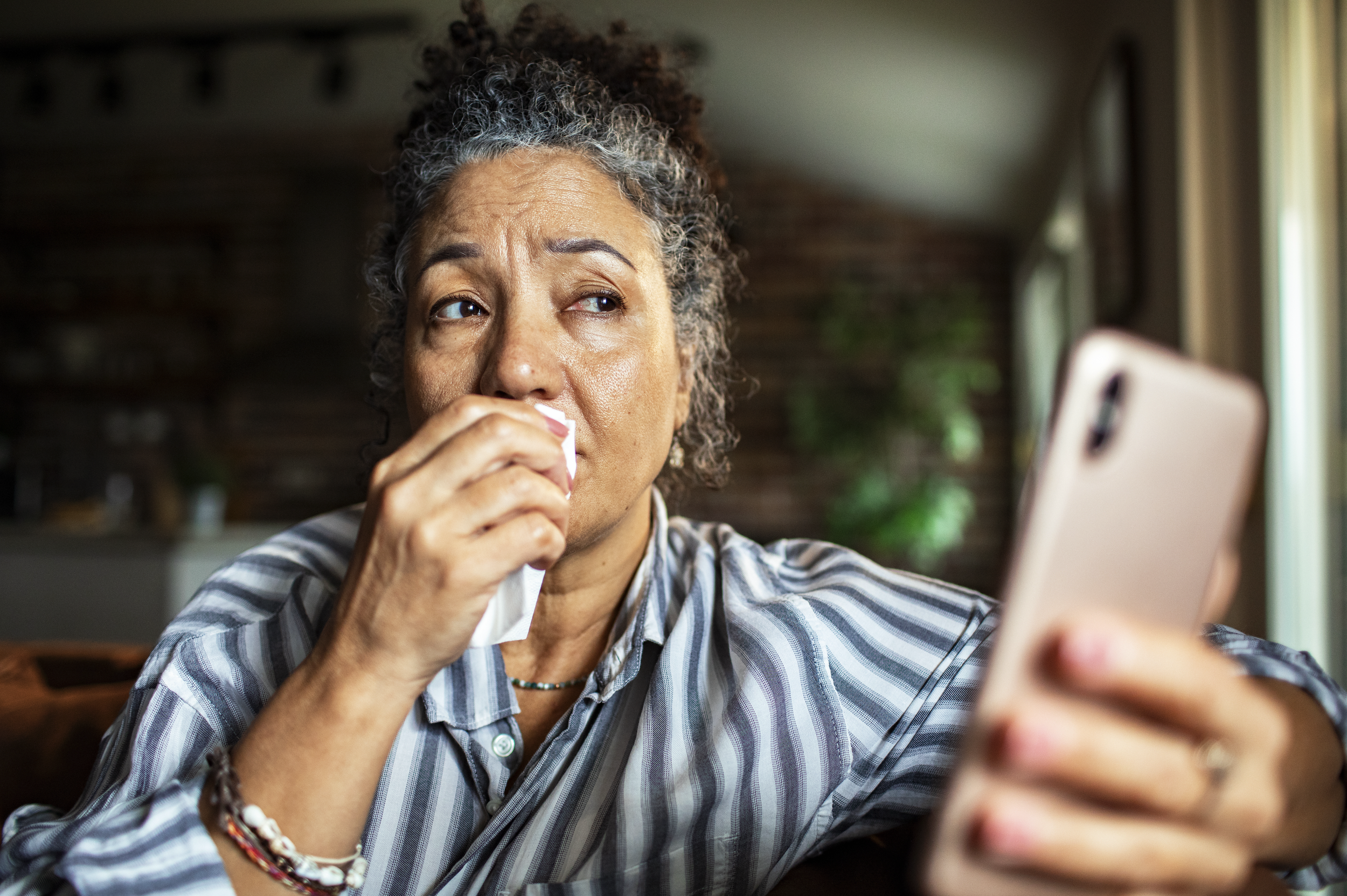 An older woman with curly gray hair is looking at her smartphone with a tissue in her nose, suggesting she might be sneezing or having a cold.
