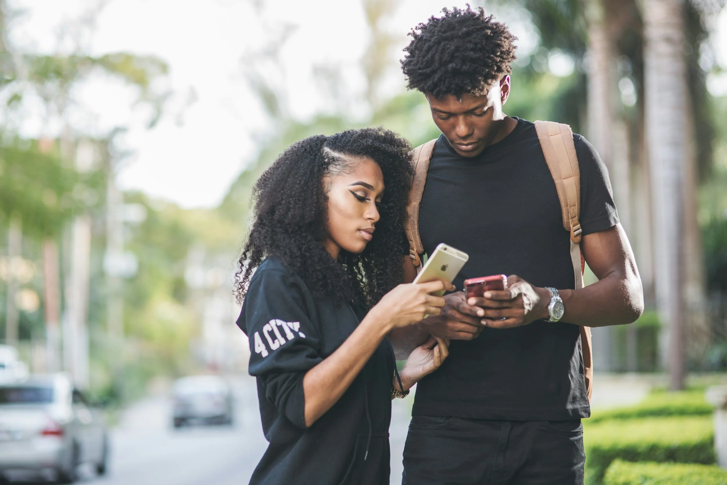 A young woman and a young man standing outdoors in a park, both looking at their smartphones. The woman has curly hair and is wearing a black top, while the man has an afro hairstyle, a black t-shirt, and a backpack.