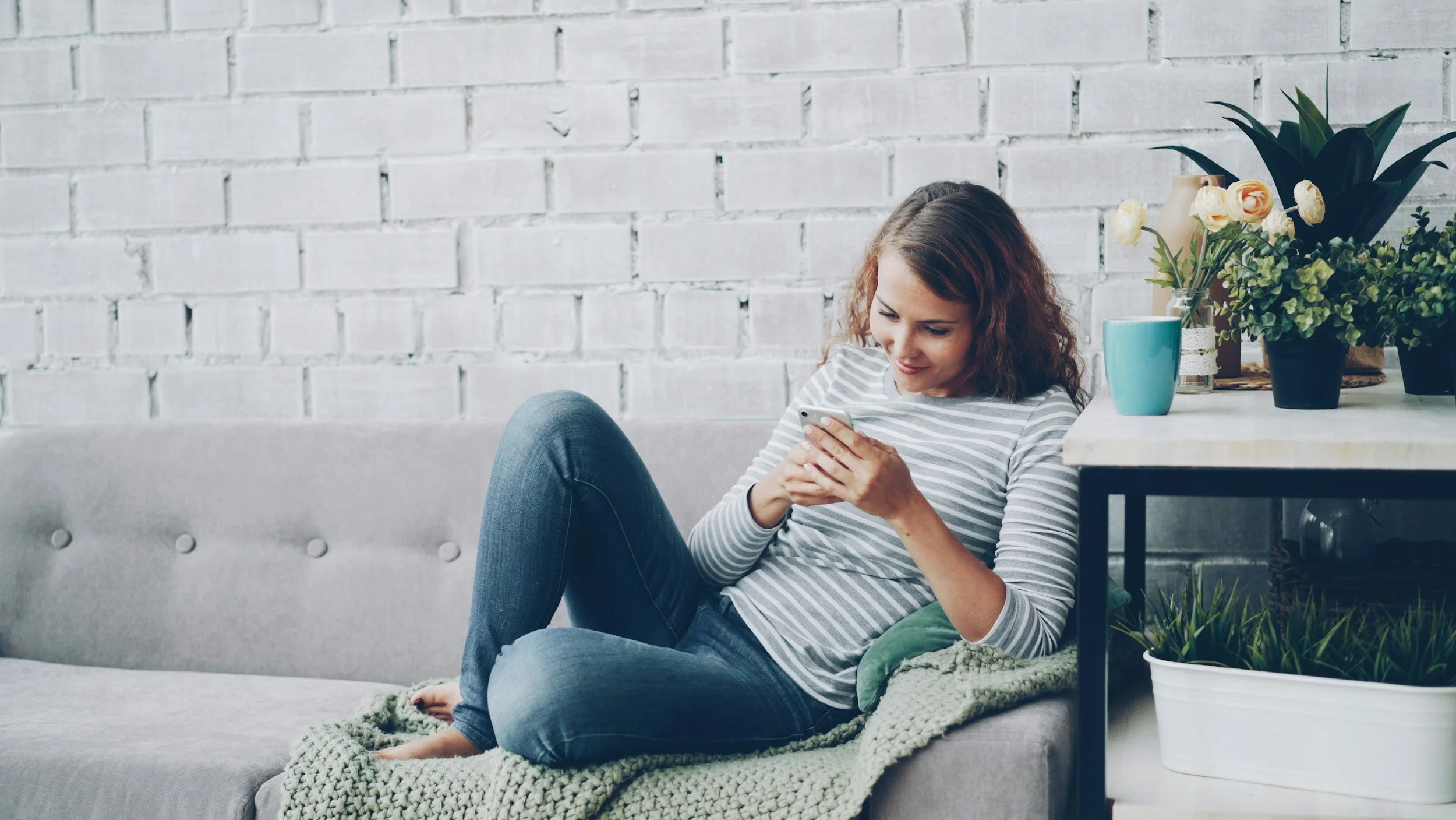 Woman with curly brown hair sitting on a gray couch, looking at her smartphone with a smile, in a room with a white brick wall, decorated with flowers and plants on a side table.