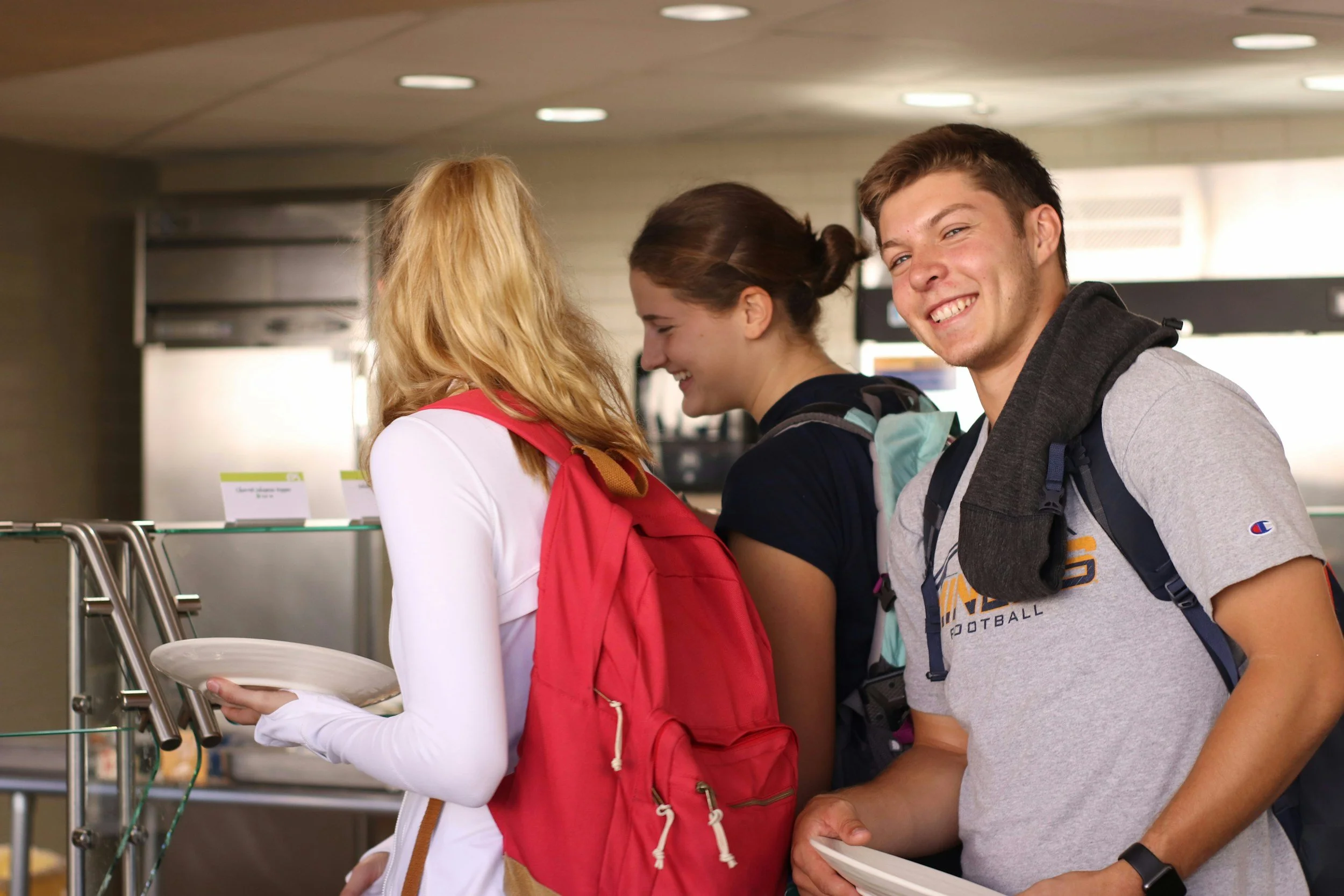 Three teenagers, two girls and a boy, standing in line at an airport.
The girl with long blonde hair wearing a white shirt and red backpack holding a white plate, smiling and looking down.
The girl with brown hair tied back, wearing a dark shirt and backpack, looking down and smiling.
The boy with short brown hair, wearing a gray T-shirt with football logo, smiling at the camera.