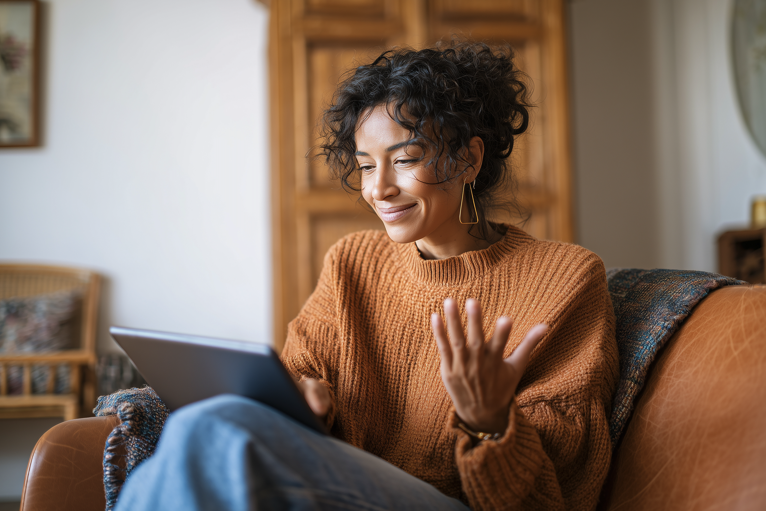 Woman sitting on a sofa using a tablet, smiling and waving her hand, in a cozy living room.