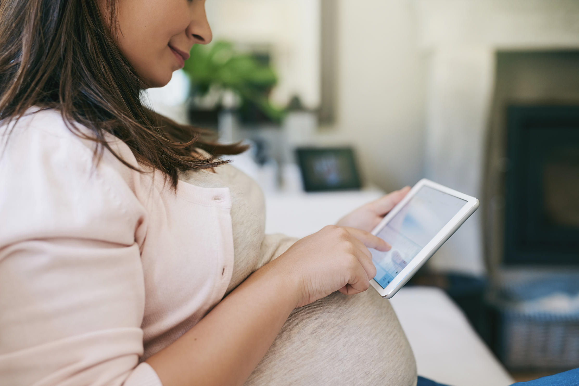 Pregnant woman sitting on a couch, using a tablet with a white case.