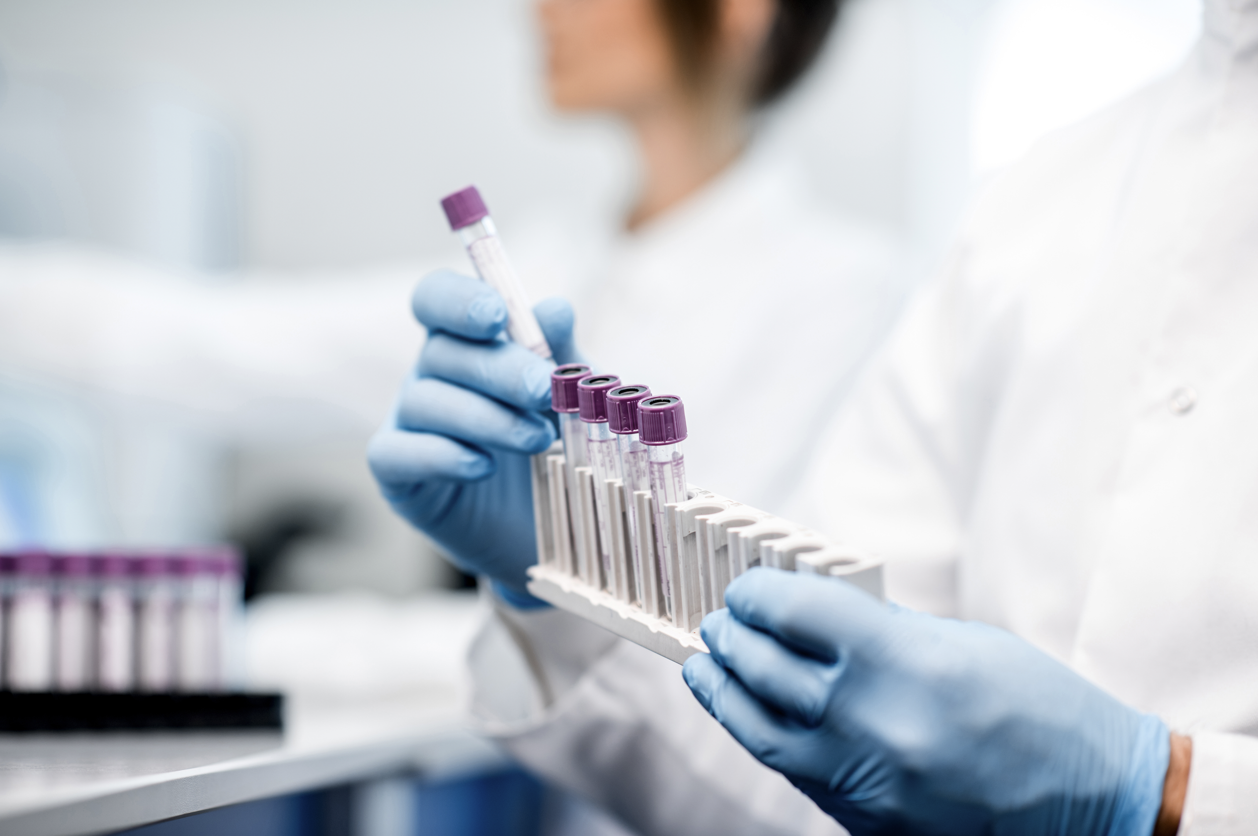 A scientist wearing blue gloves holding a rack of test tubes filled with a pink liquid in a laboratory.