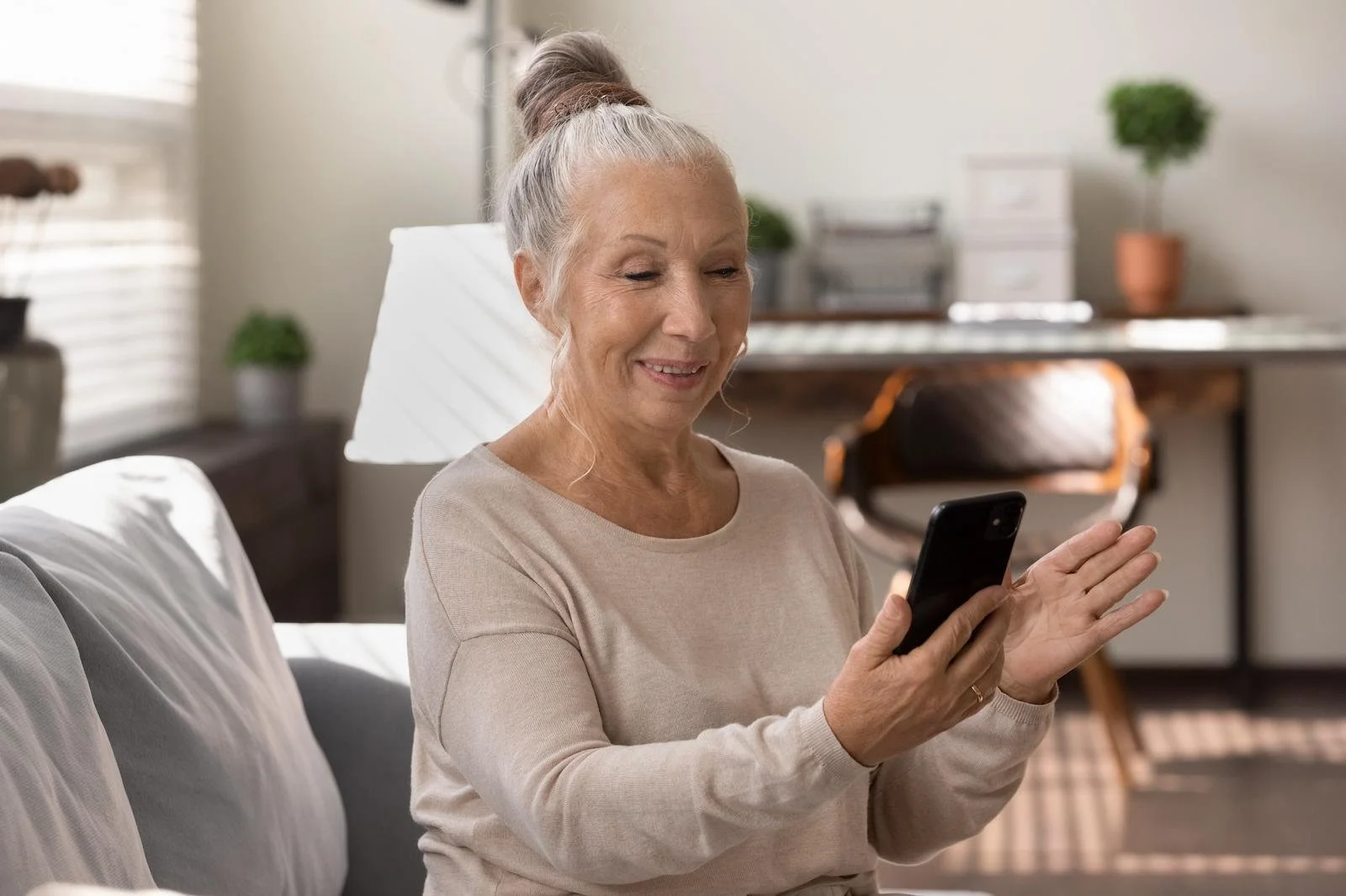 An elderly woman with gray hair in a bun smiling and waving while using a smartphone in her living room.