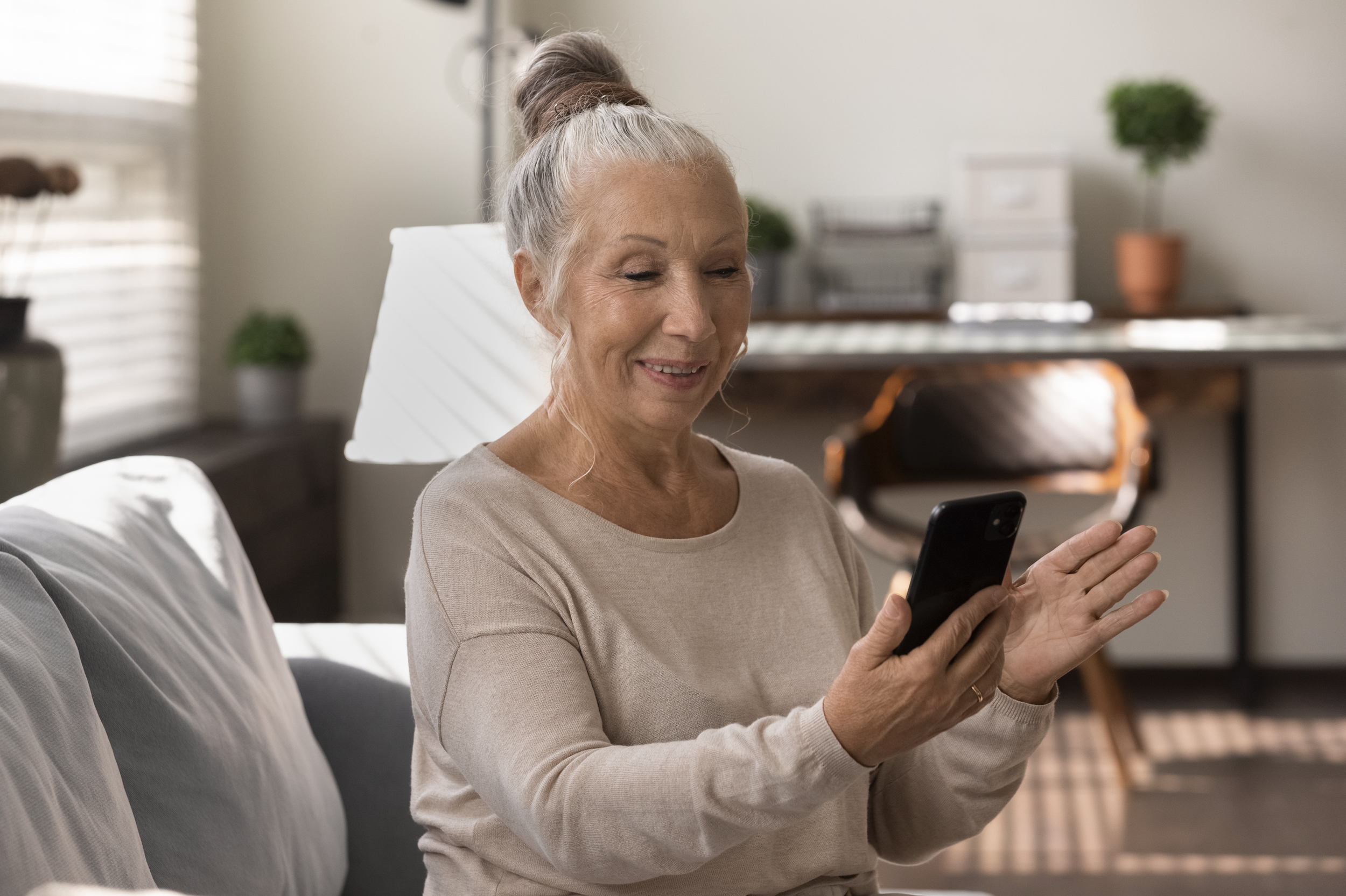 An elderly woman with gray hair in a messy bun smiling while looking at her smartphone in a bright living room.