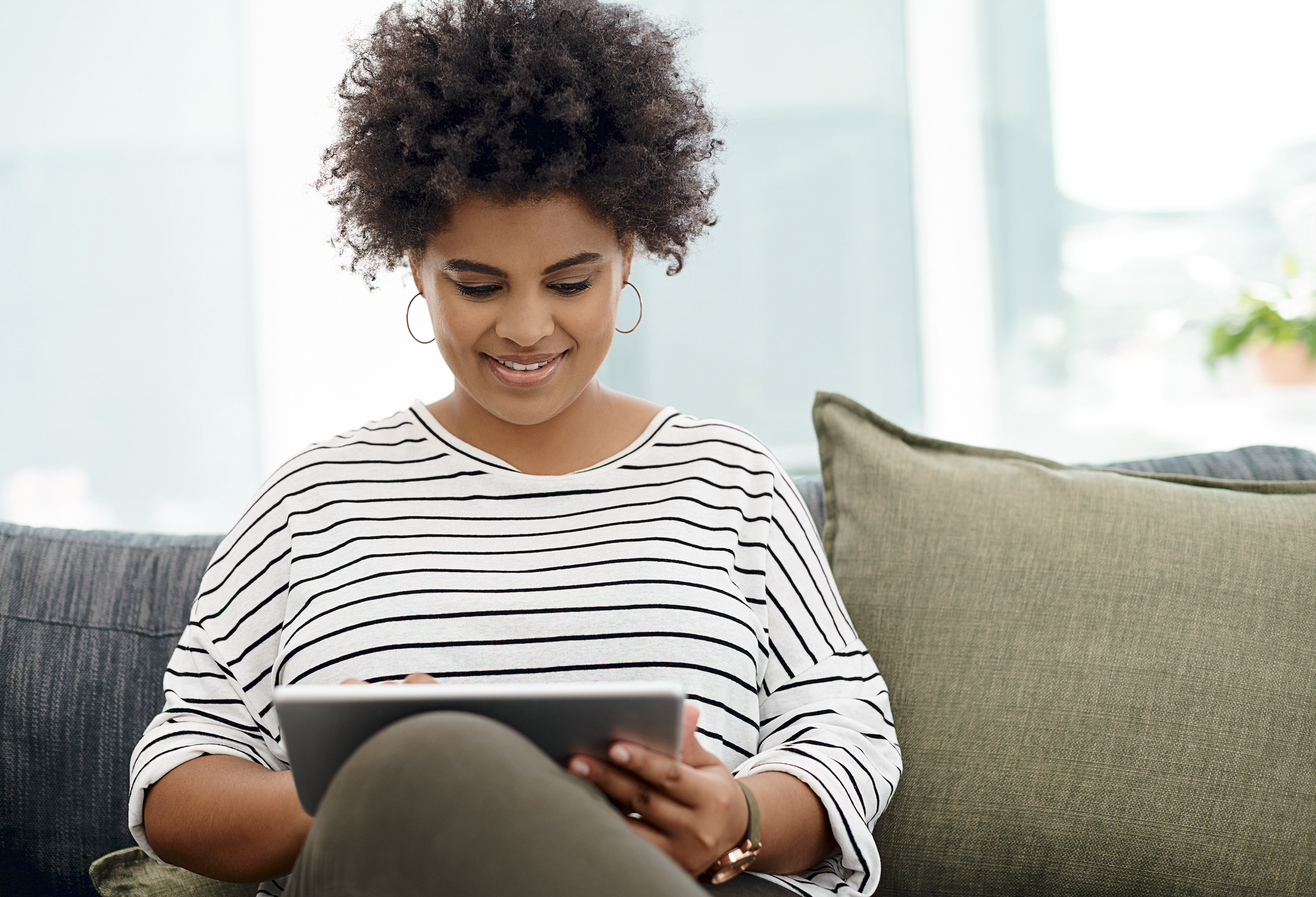 A woman with curly hair and hoop earrings smiling while using a tablet, sitting on a sofa with green and gray cushions in a bright room.