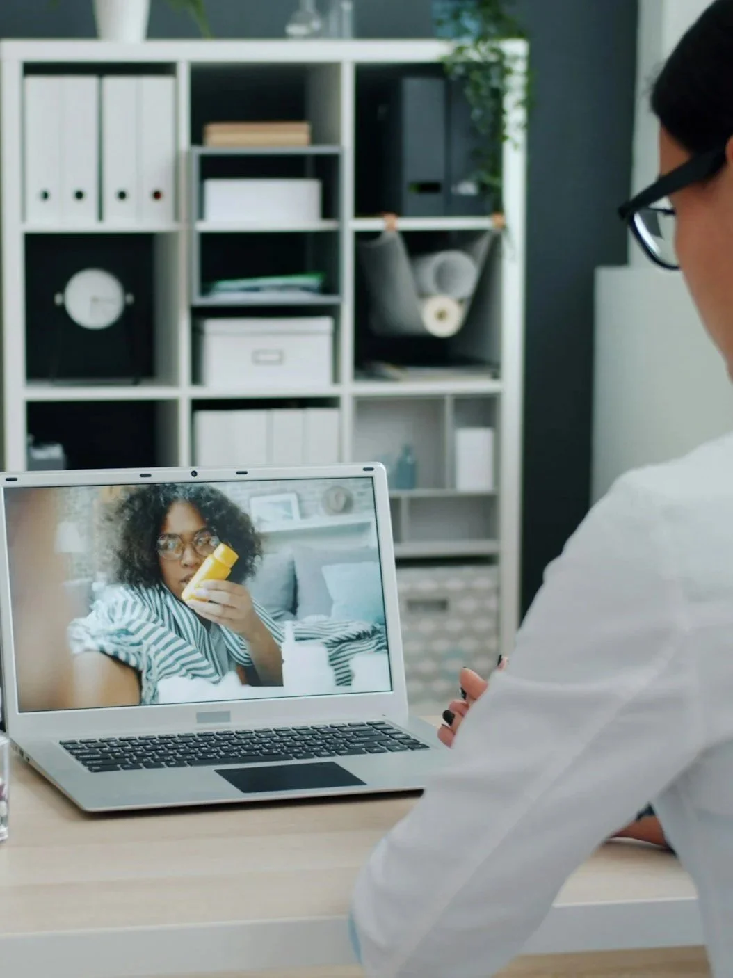 A woman sits at a desk looking at a laptop, on the screen is a woman with curly hair and glasses holding a yellow bottle, in a home office with shelves in the background.