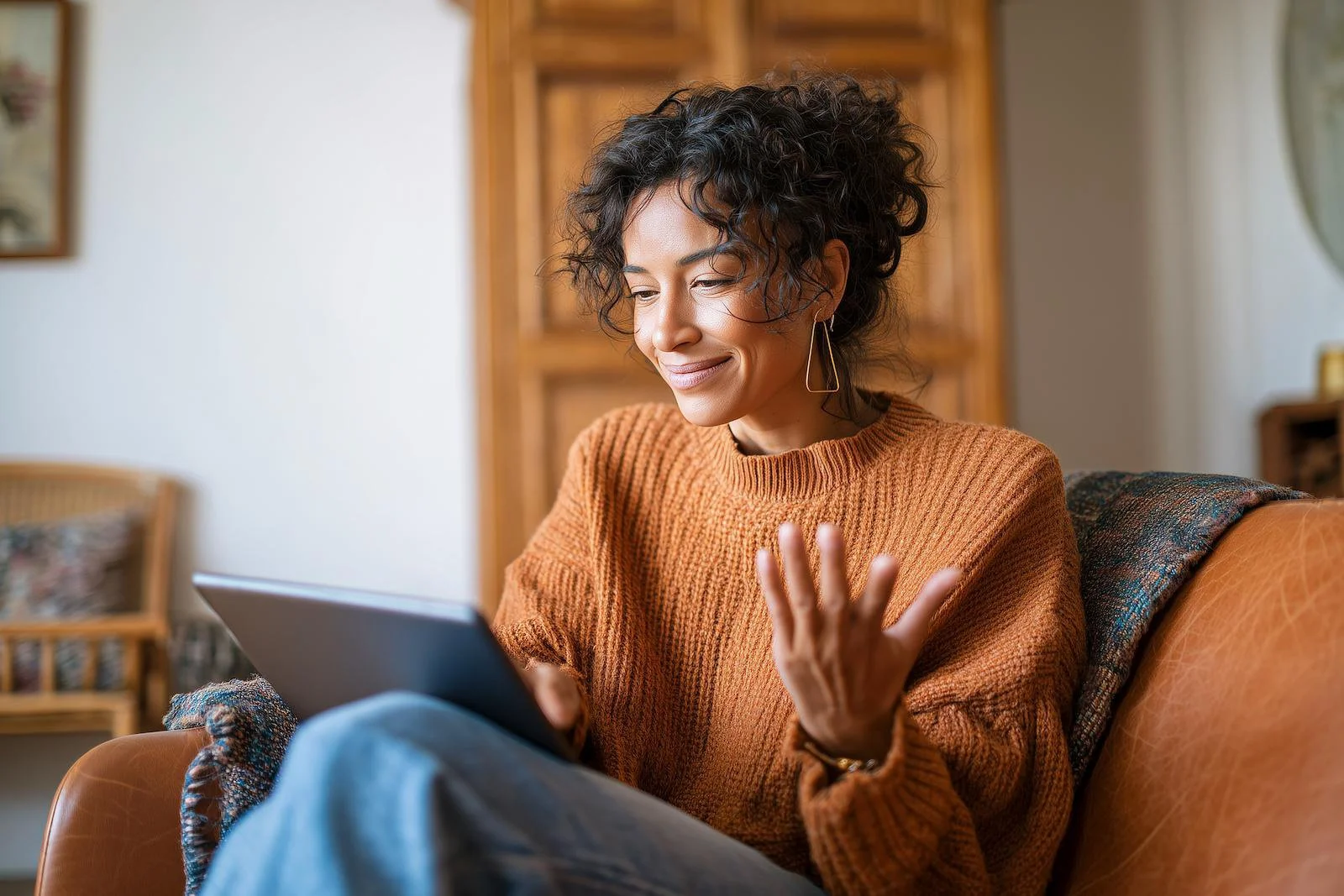 A woman with curly hair wearing an orange sweater and earrings sitting on a couch, looking at her tablet with a smile