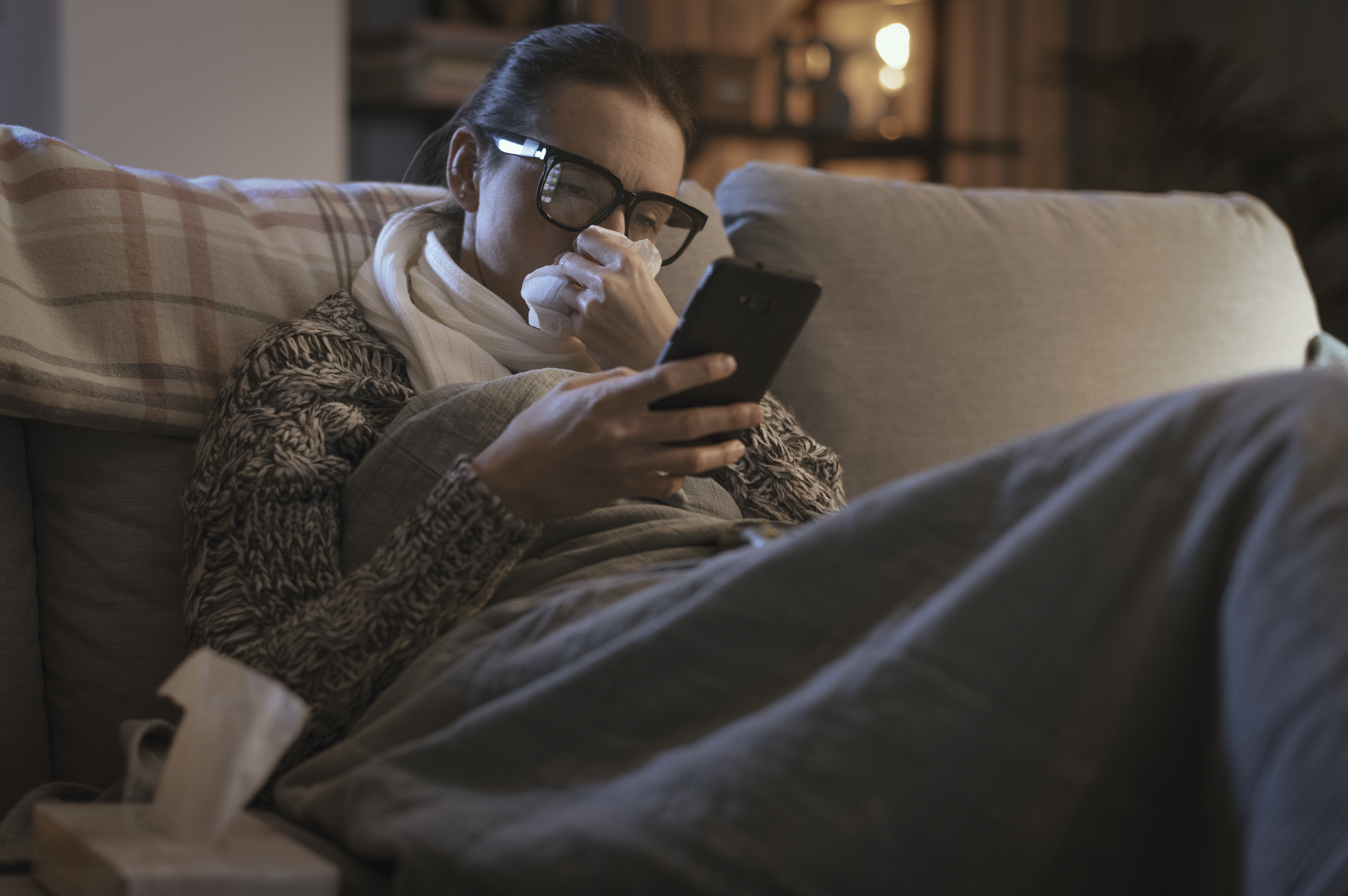 A woman with glasses and a scarf sitting on a couch, holding a tissue to her nose and looking at her phone, with a box of tissues on the table in front of her.