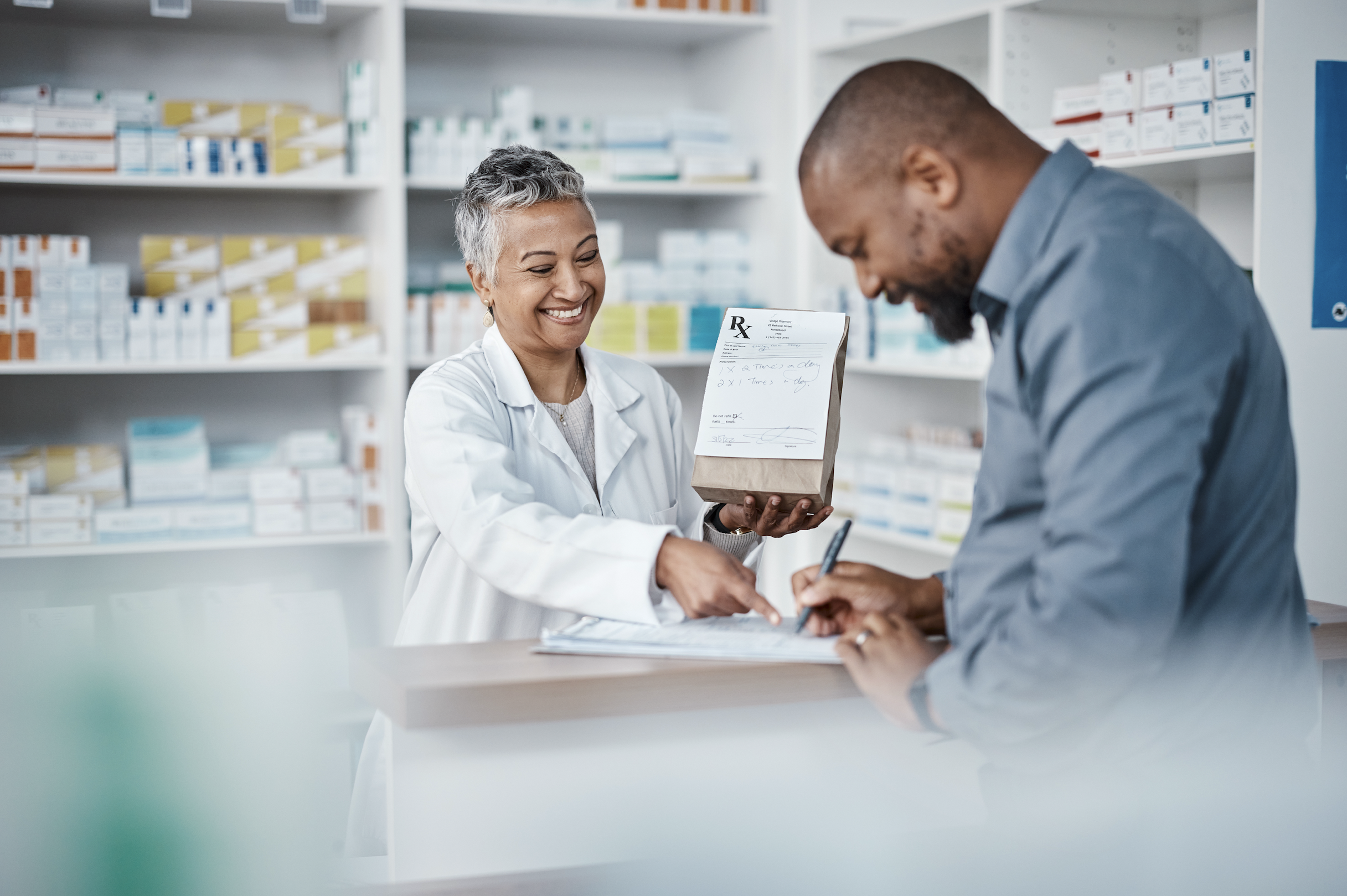 A pharmacist handing a paper bag to a customer at a pharmacy counter with shelves of medicines in the background.
