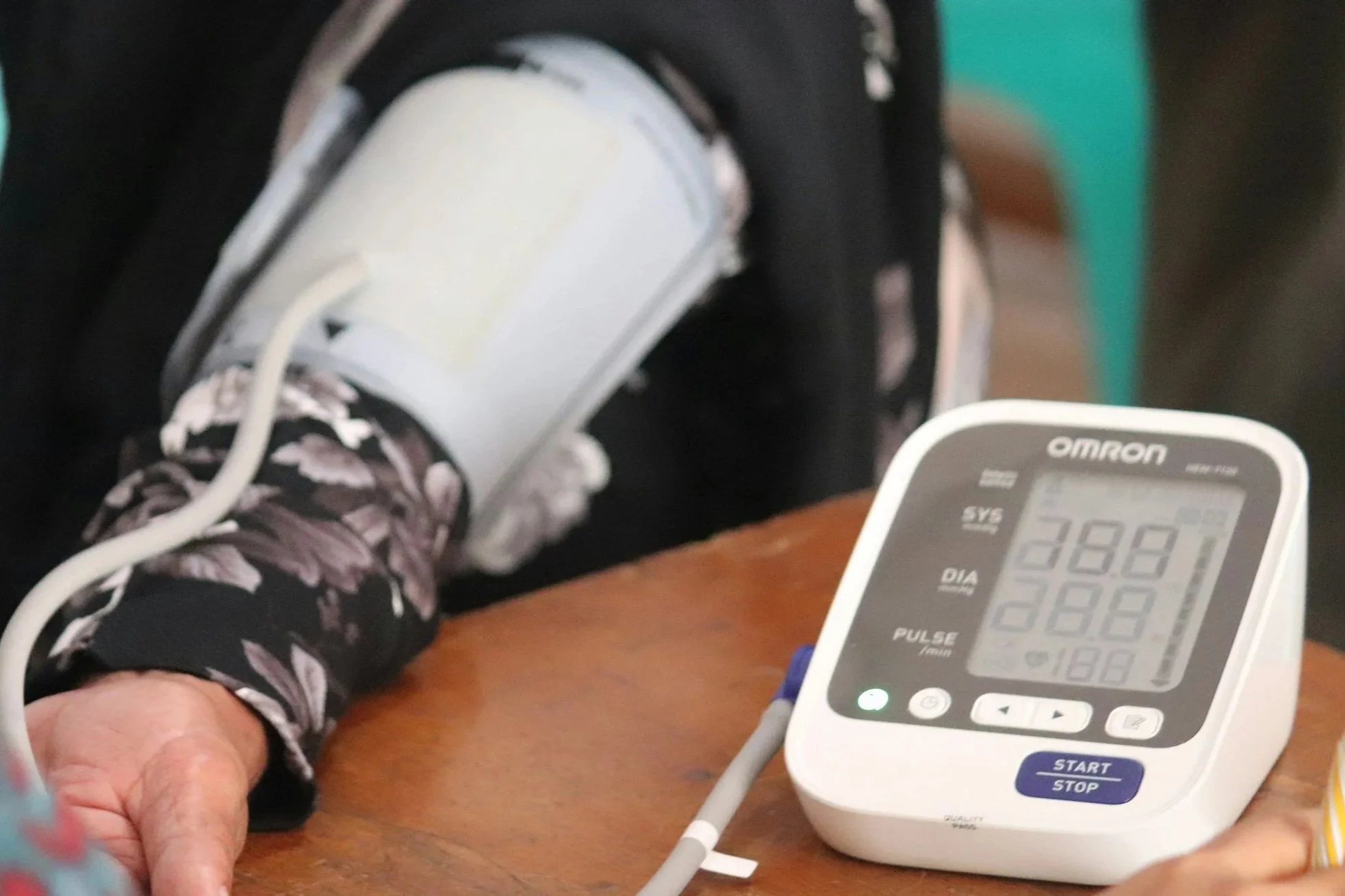 Close-up of a person's arm with a cuff on their upper arm, a digital blood pressure monitor displaying 138/88, and a hand resting on a wooden surface.