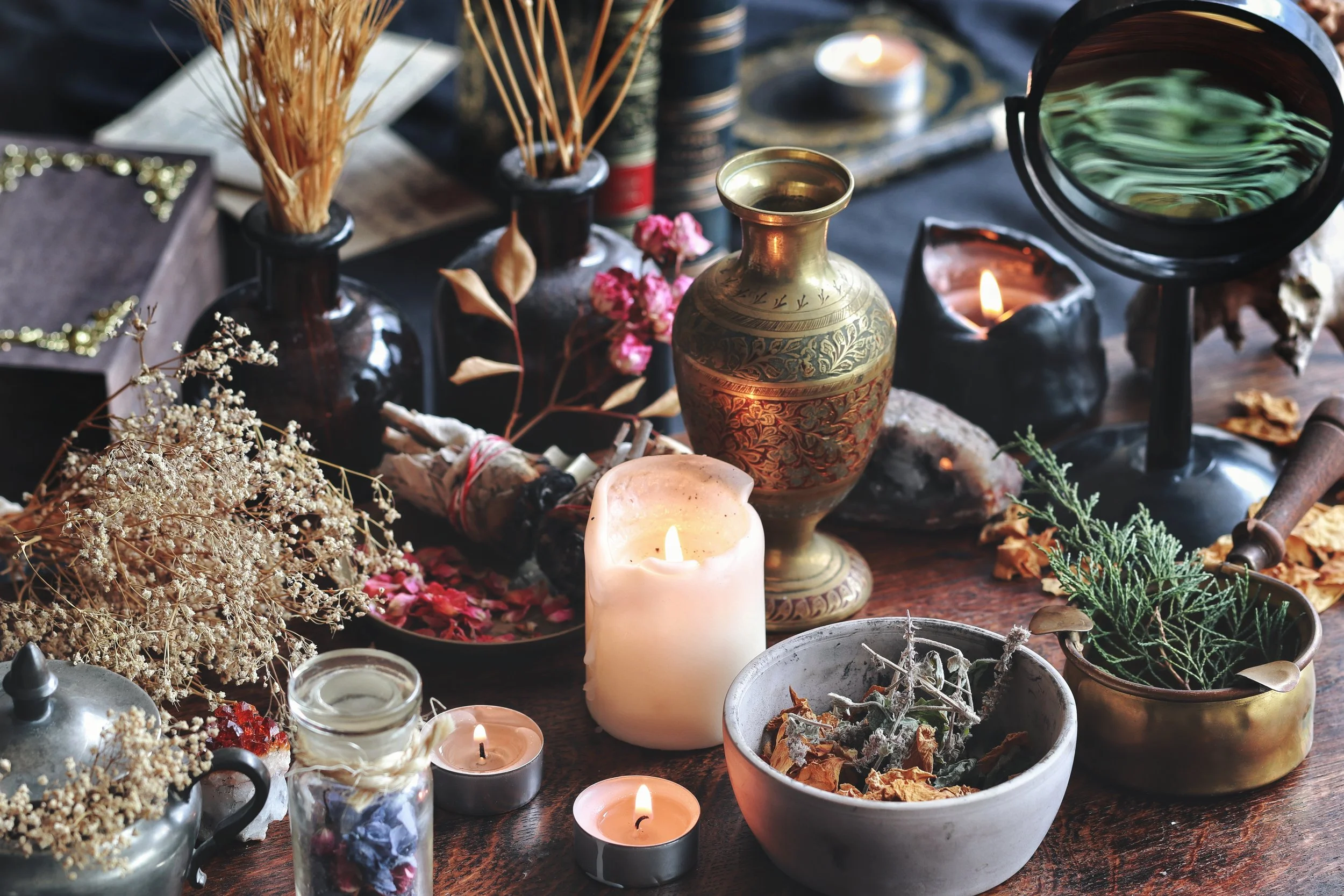 A table with various decorative items including candles, dried flowers, a brass vase, and small jars and bowls, arranged for a spiritual or ritual setting.