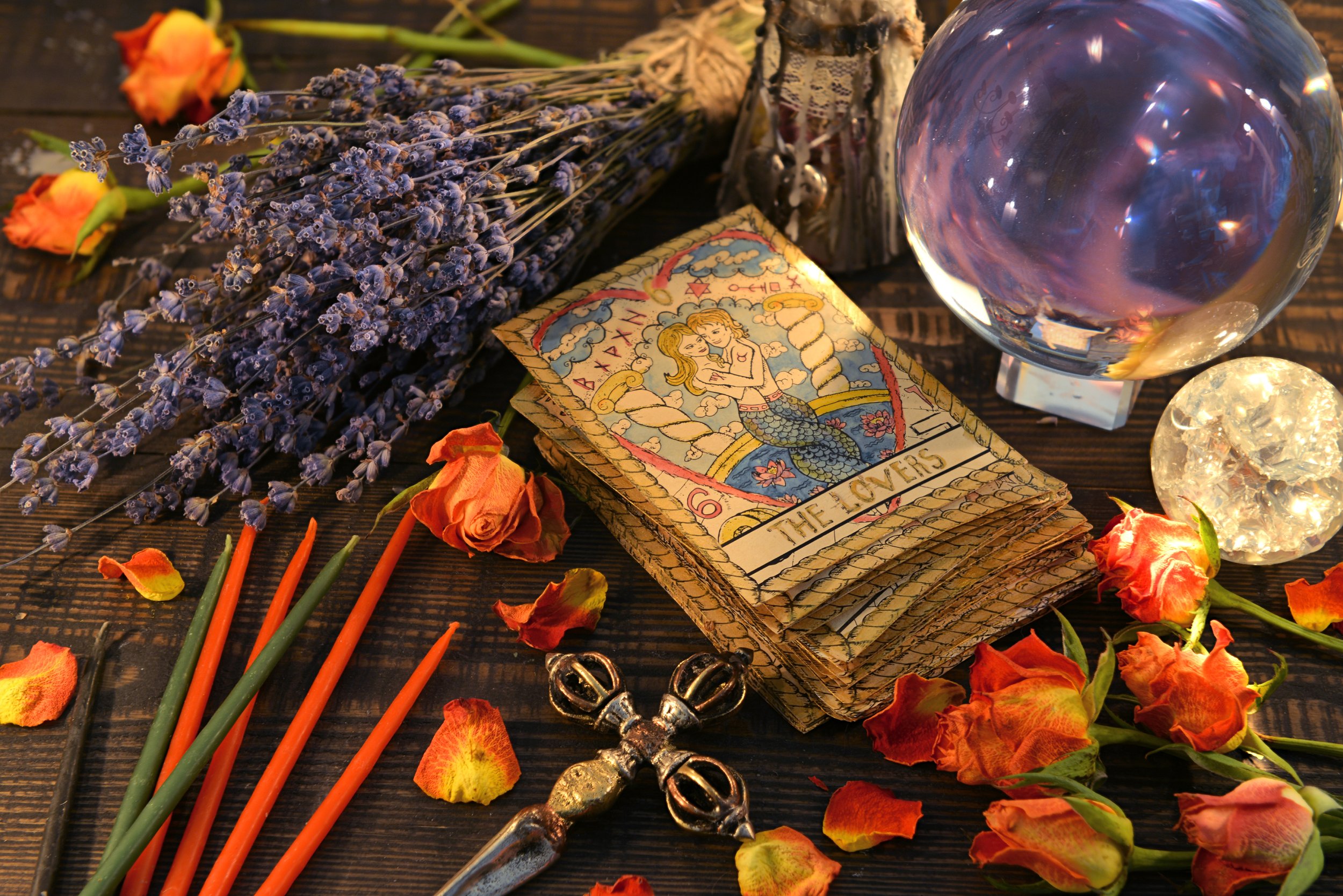 A ritual setup on a dark wooden surface includes dried lavender, burnt orange roses, some candles, a crystal ball, a tarot card, and metal charms, surrounded by scattered dried flower petals.