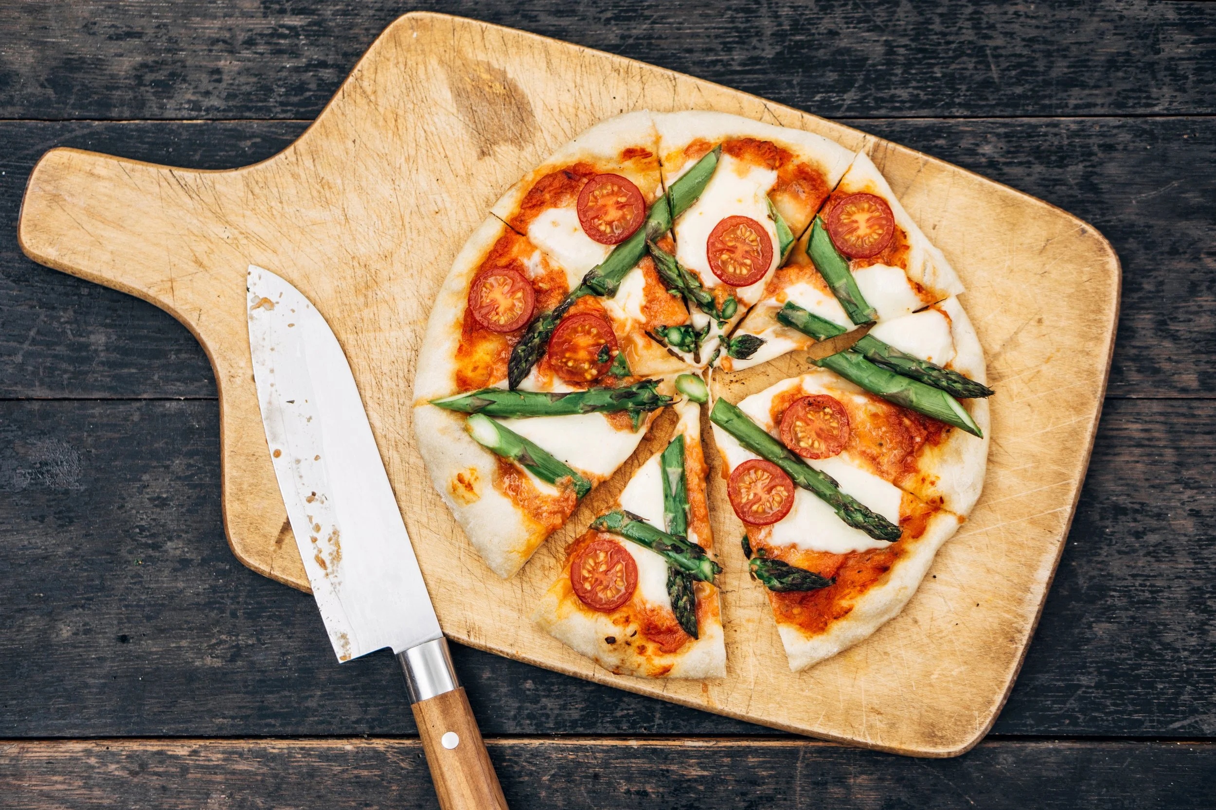 A partially sliced pizza with cherry tomatoes and green asparagus on a wooden paddle, on a dark wooden surface.