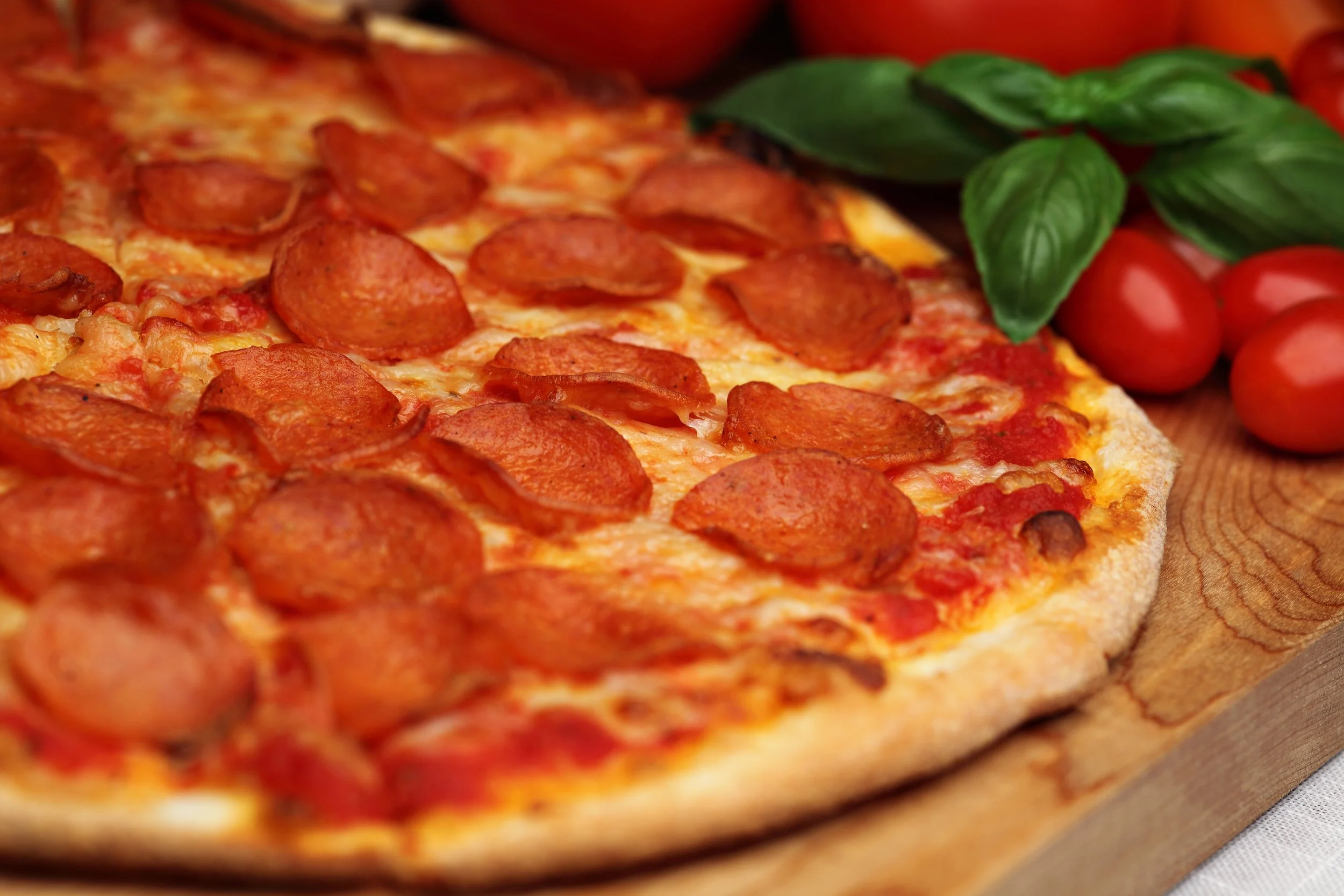 Close-up of a pepperoni pizza on a wooden serving board with cherry tomatoes and basil leaves in the background.