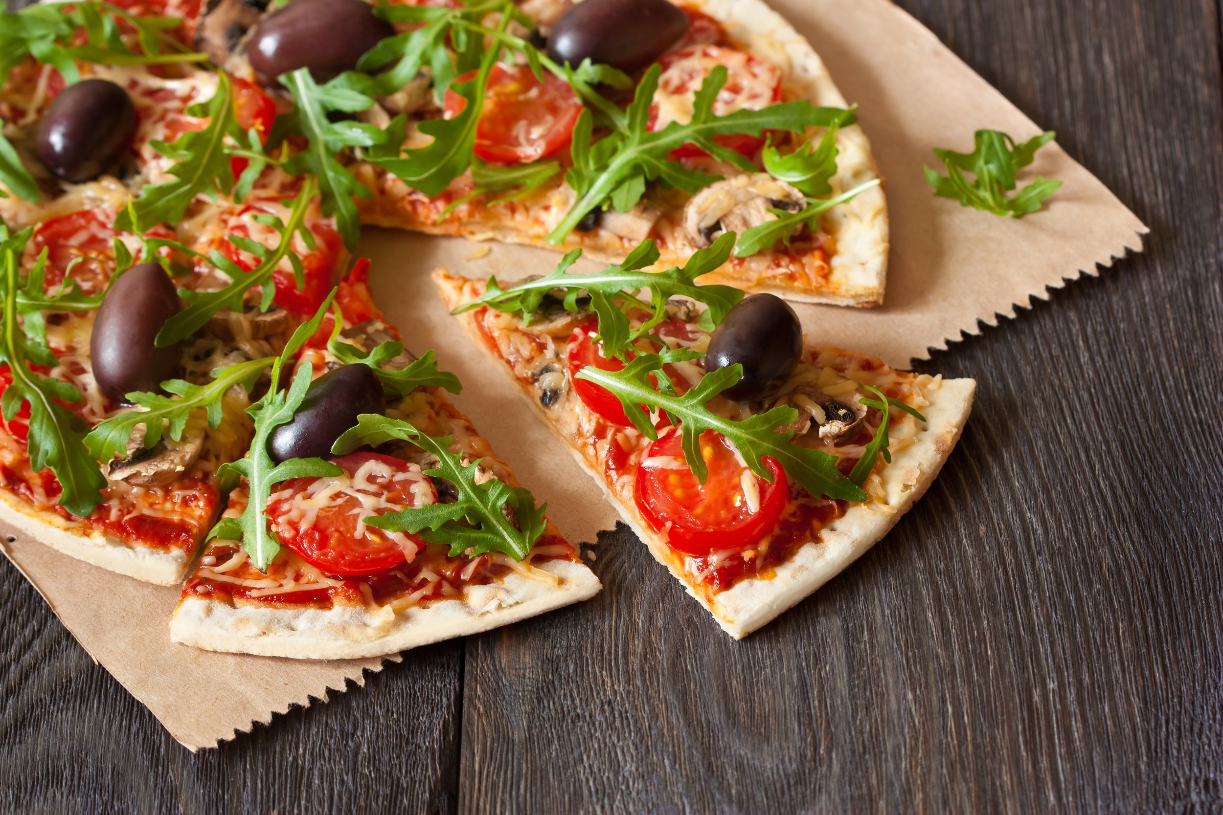 Personal-sized vegetable pizza with cherry tomatoes, black olives, arugula, and shredded cheese on a wooden surface.