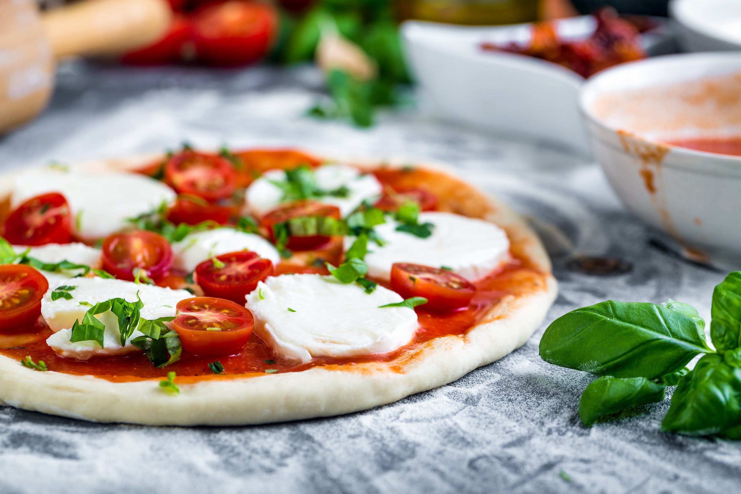 Fresh Margherita pizza with cherry tomatoes, mozzarella, and basil on parchment paper, with bowls of sauce in the background.