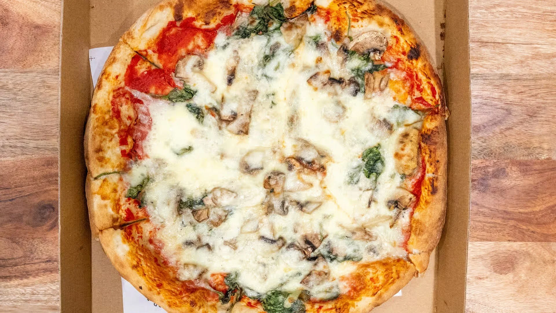 Cheese pizza with mushrooms, spinach, and tomato slices in a cardboard box on a wooden table.
