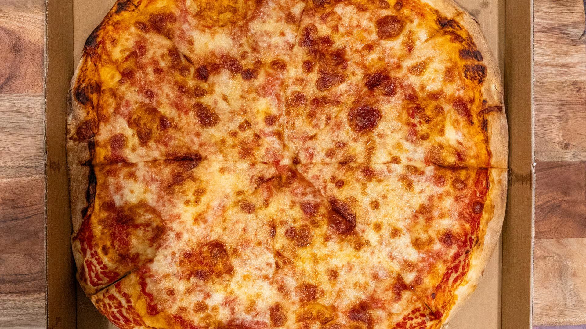 A large cheese pizza with a golden crust and melted cheese in a box on a wooden surface.