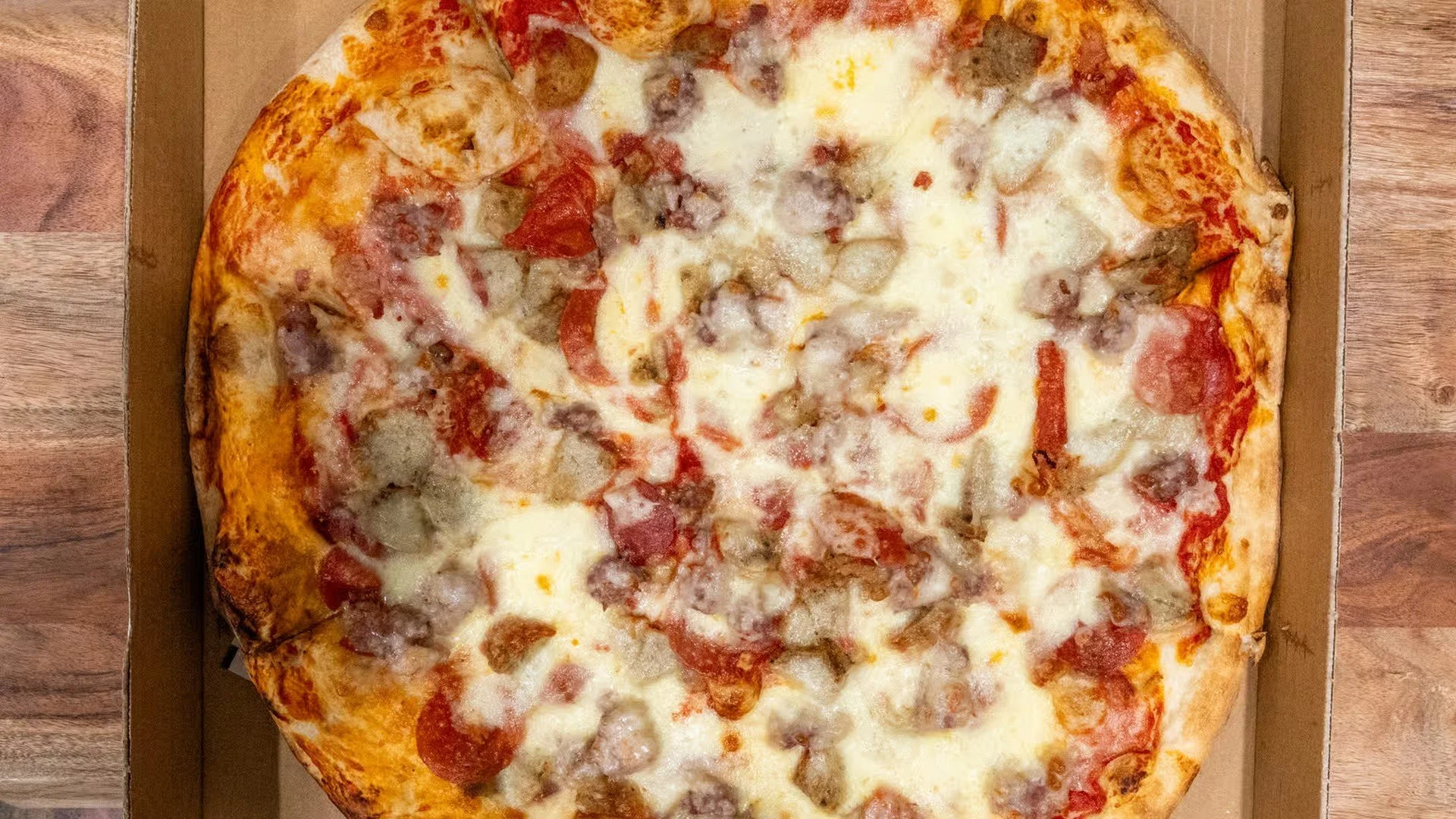 A whole cheese pizza with pepperoni, sausage, and bacon, sitting in an open pizza box on a wooden surface.