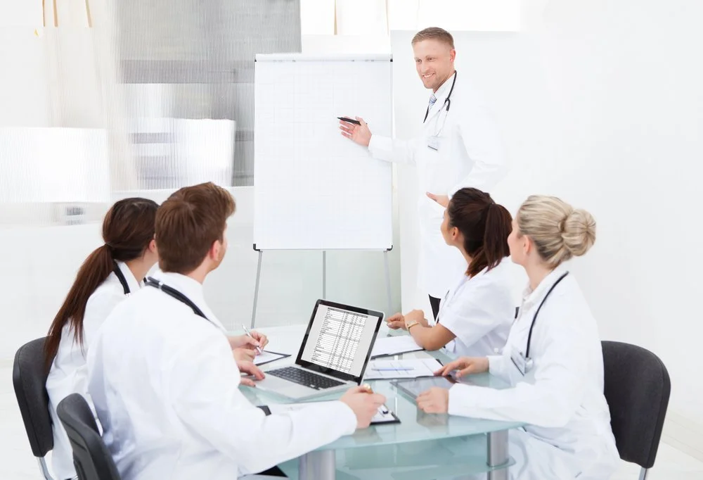 Medical team in a meeting room, with a doctor standing by a whiteboard and five team members seated at a table with laptops and papers.