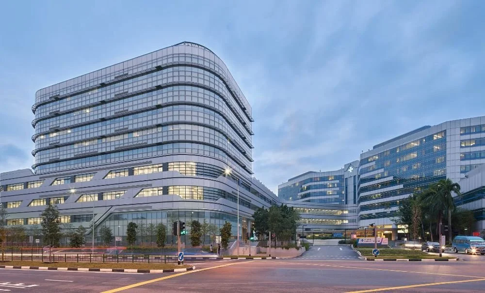 Modern hospital buildings with glass facades on a city street at dusk, with streetlights and some cars visible.
