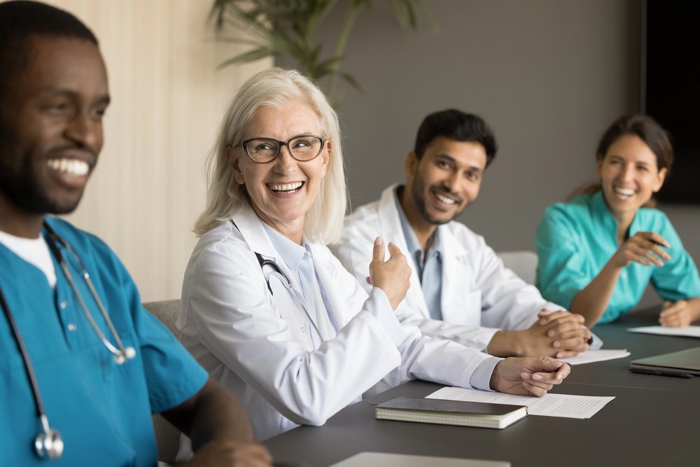Medical professionals, including doctors and nurses, are sitting at a conference table, smiling and engaging in conversation during a meeting in a hospital or clinic setting.
