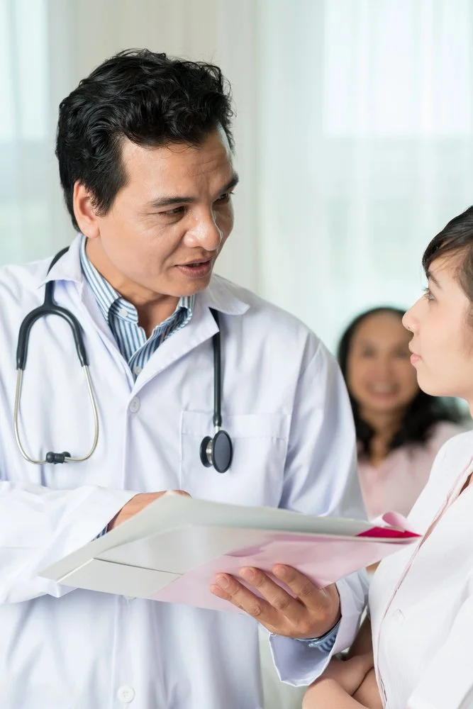 A male doctor with a stethoscope around his neck talking to a female patient in a medical office, while another woman stands in the background.