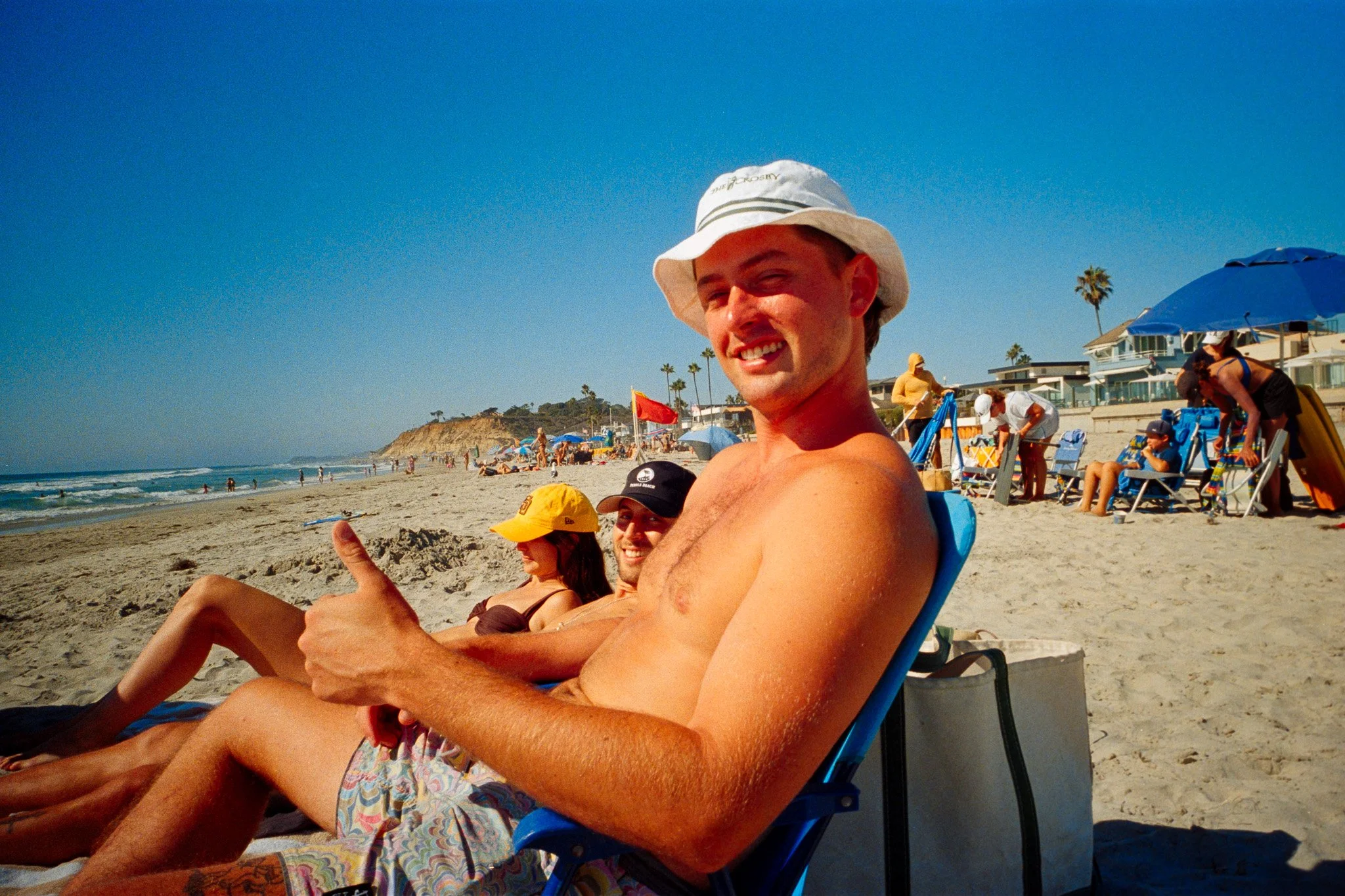 A group of people relaxing on a sandy beach with blue umbrellas, palm trees, and beach houses in the background. One man is smiling and giving a thumbs-up, wearing a white bucket hat.