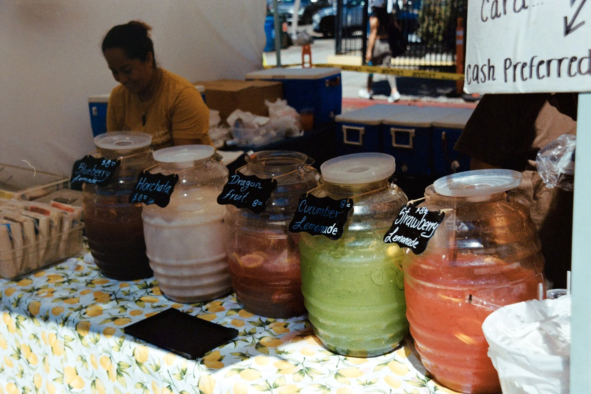 A woman behind a stand with large glass jars of colorful beverages labeled with black chalkboard signs, at an outdoor market with a yellow caution tape and pedestrians in the background.
