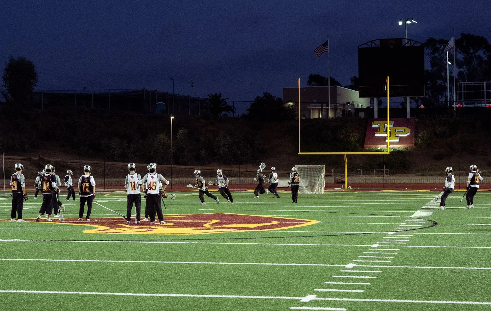 A group of people wearing sports gear on a football field at night, with stadium lights and an American flag in the background.