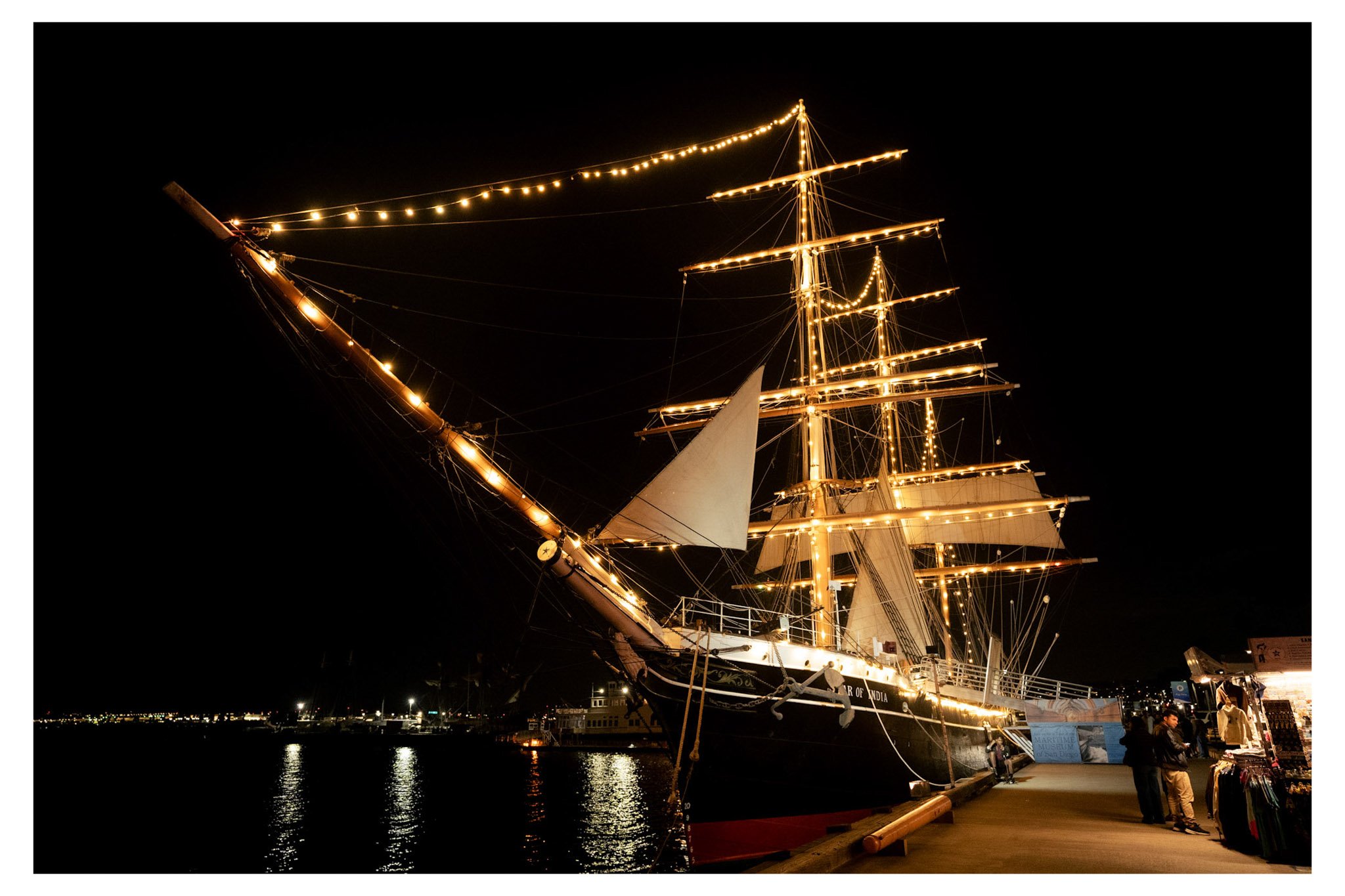 A tall ship decorated with string lights docked at a harbor at night, with reflections on the water and illuminated cityscape in the background.