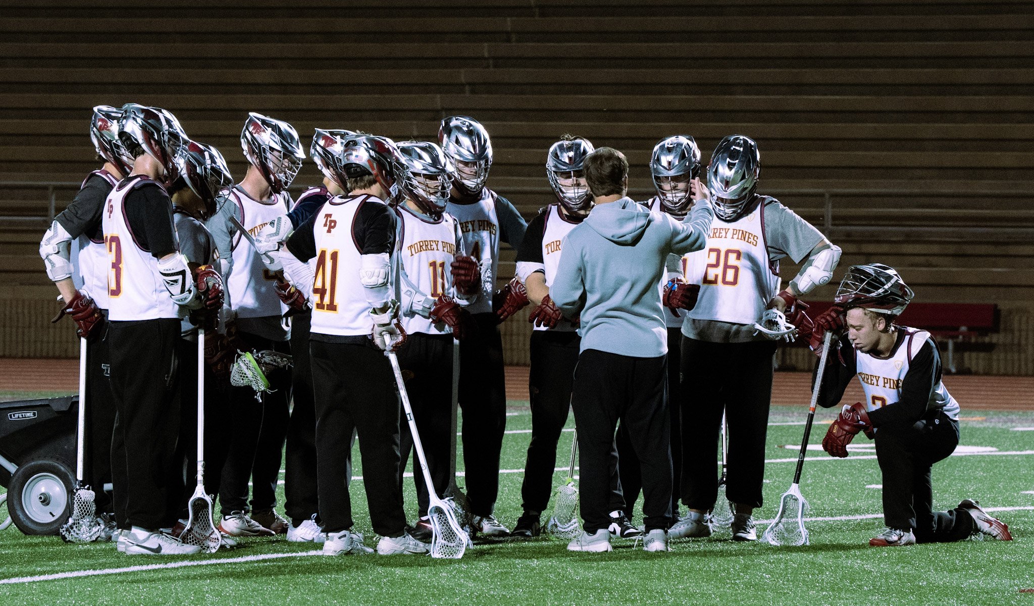 A group of lacrosse players wearing helmets and jerseys with 'Torrey Pines' written on them, gathered around their coach on a field at night.