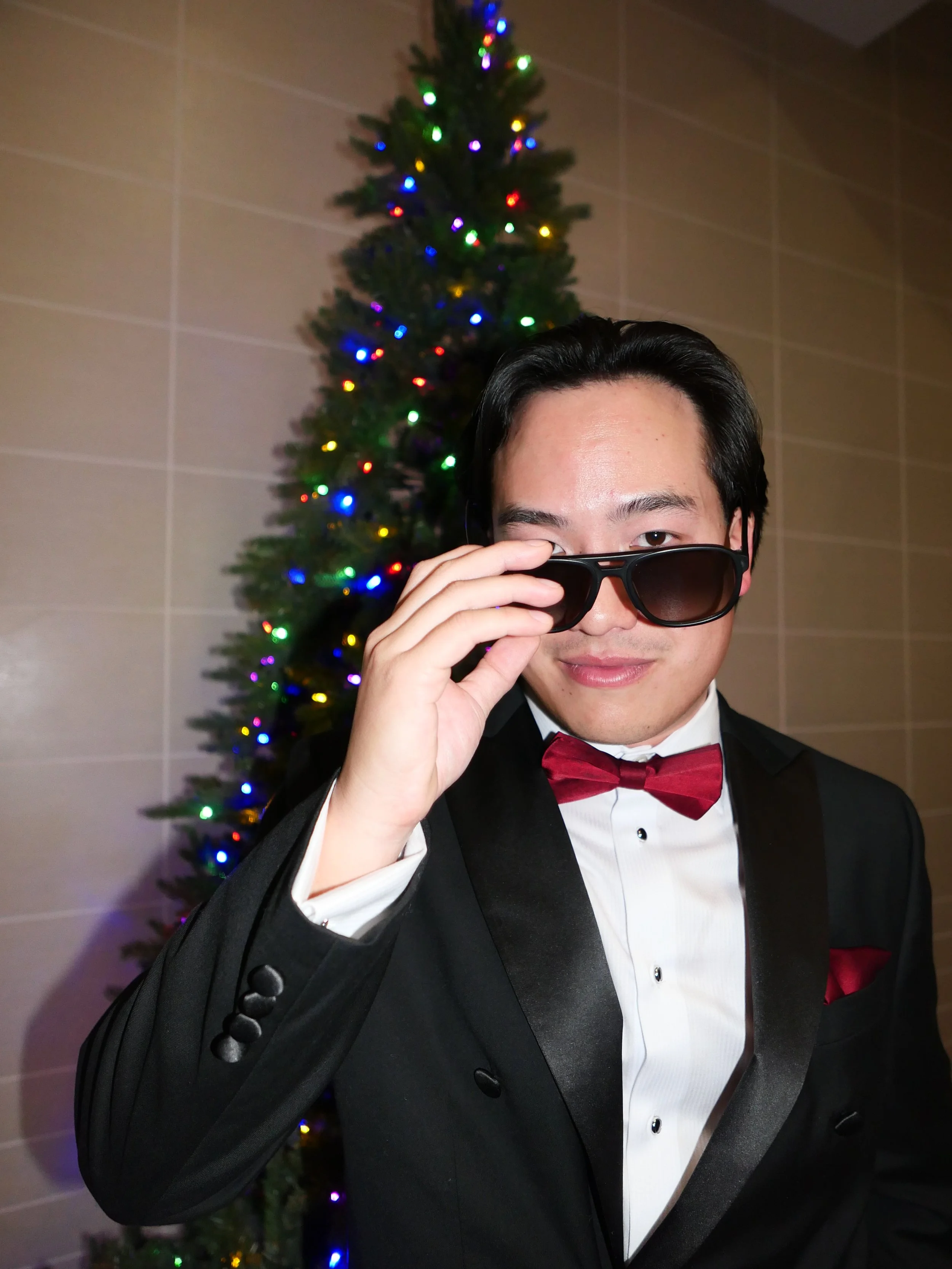 A man dressed in a formal black tuxedo with a white shirt and red bow tie, adjusting his sunglasses, standing in front of a decorated Christmas tree with colorful lights.