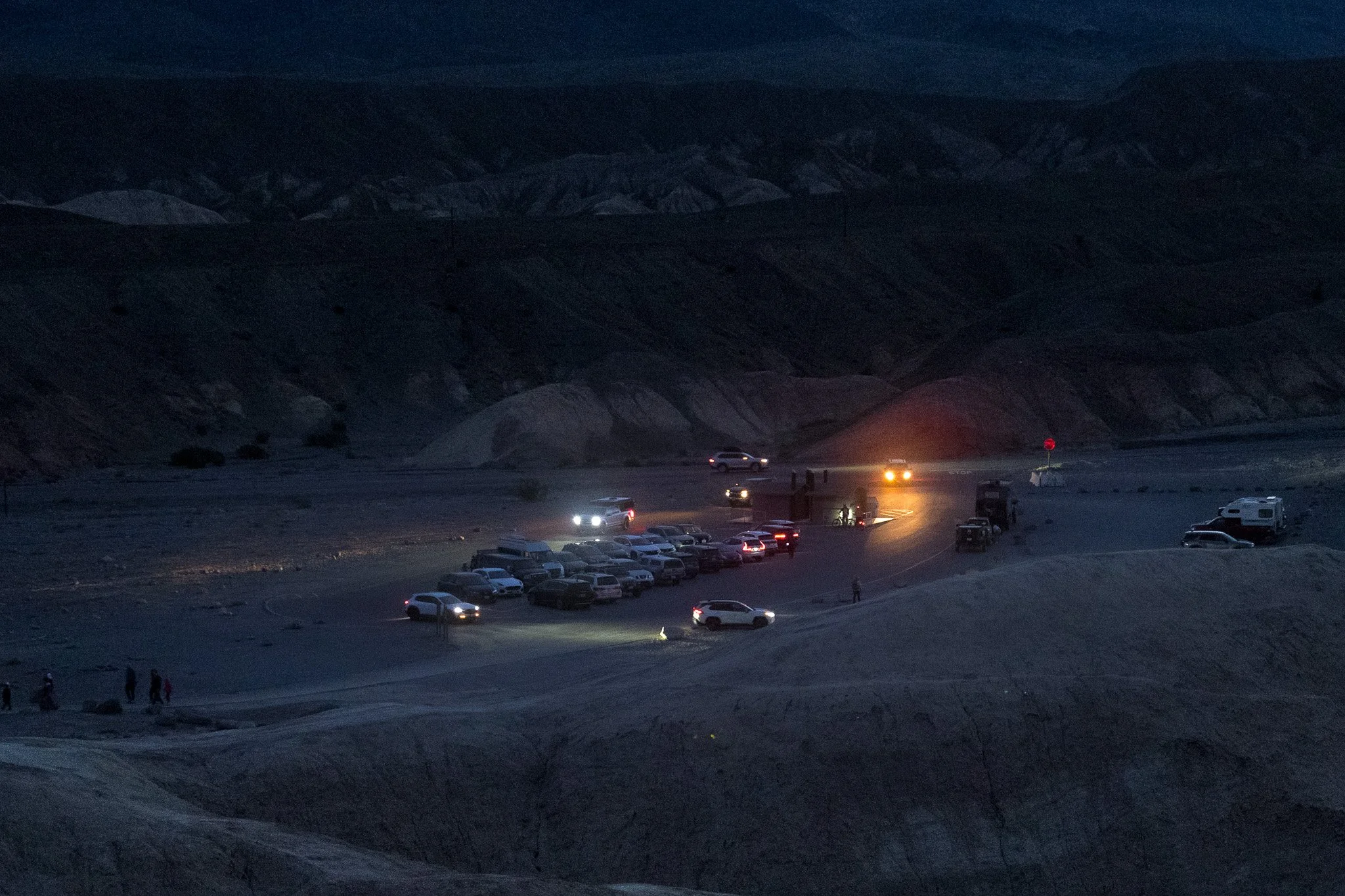 A nighttime scene of a parking lot in a desert area, with mountains in the background. Several cars are parked, and a few vehicles have their lights on. There are a few people walking around, and some structures and equipment are visible near the parked cars. The sky is dark with no visible stars or moon.